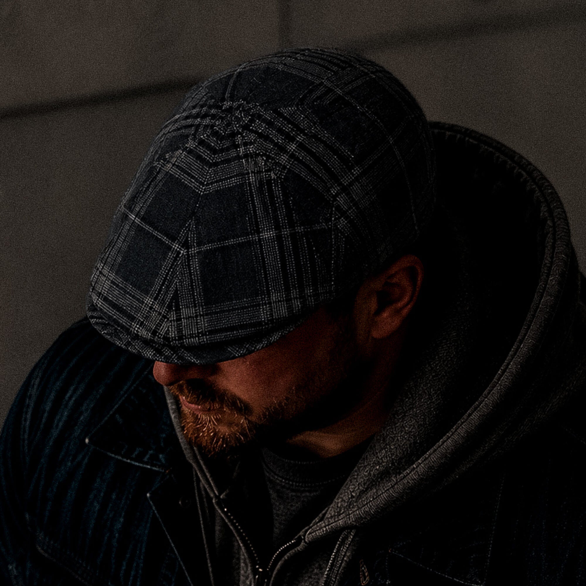 A man in the Glenwood Grove jacket by Goorin Bros., his face partially shadowed, head slightly bowed beneath the hoodie, wears a classic flatcap.