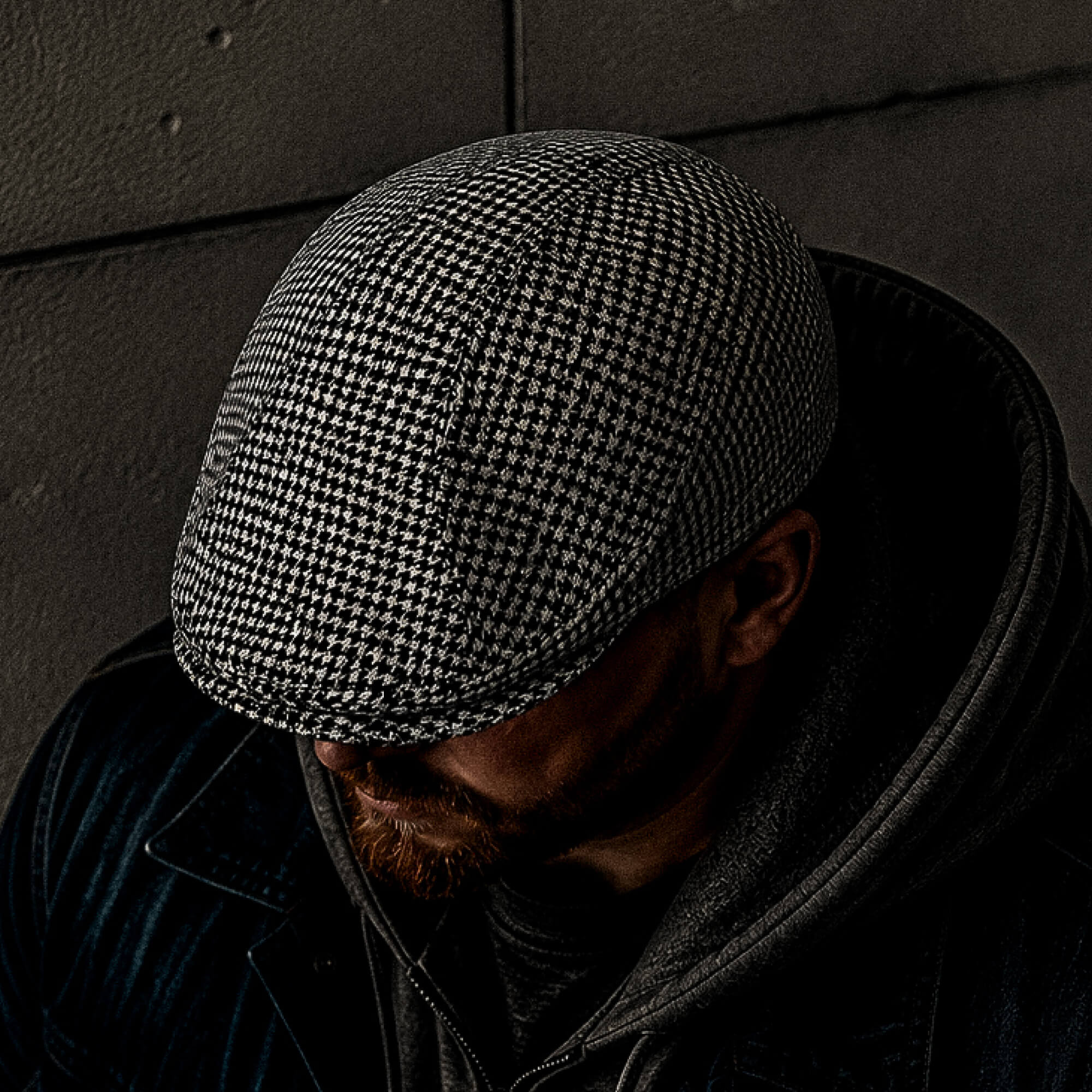 Wearing a Goorin Bros. Norwood classic flatcap in black and white houndstooth, a person in a dark jacket and hoodie bows their head to partially obscure their face.