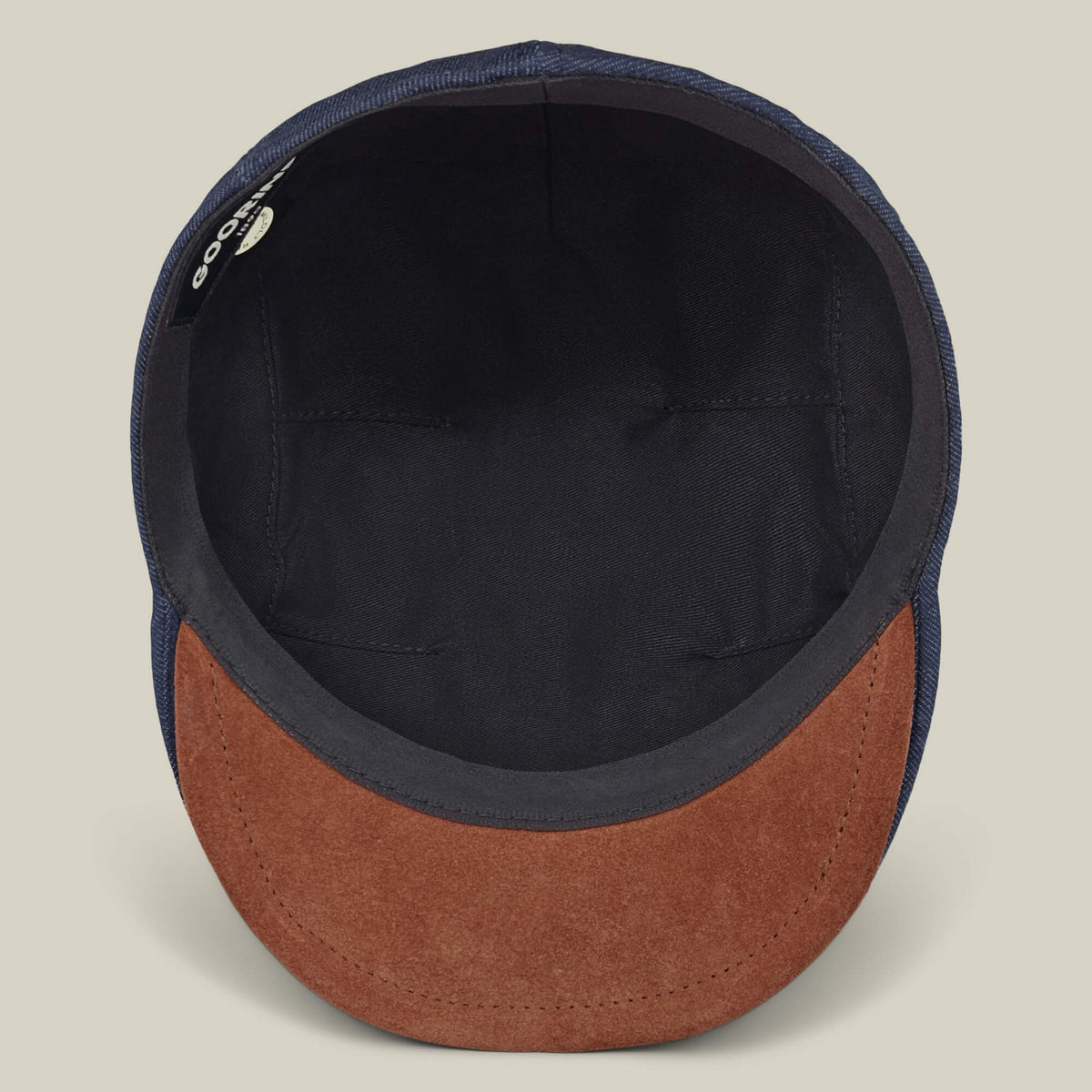 The Goorin Bros. Ashfield flat cap, hand sewn in the USA, is shown from above, highlighting its black interior lining, brown suede brim, and a small fabric brand tag on the inside.