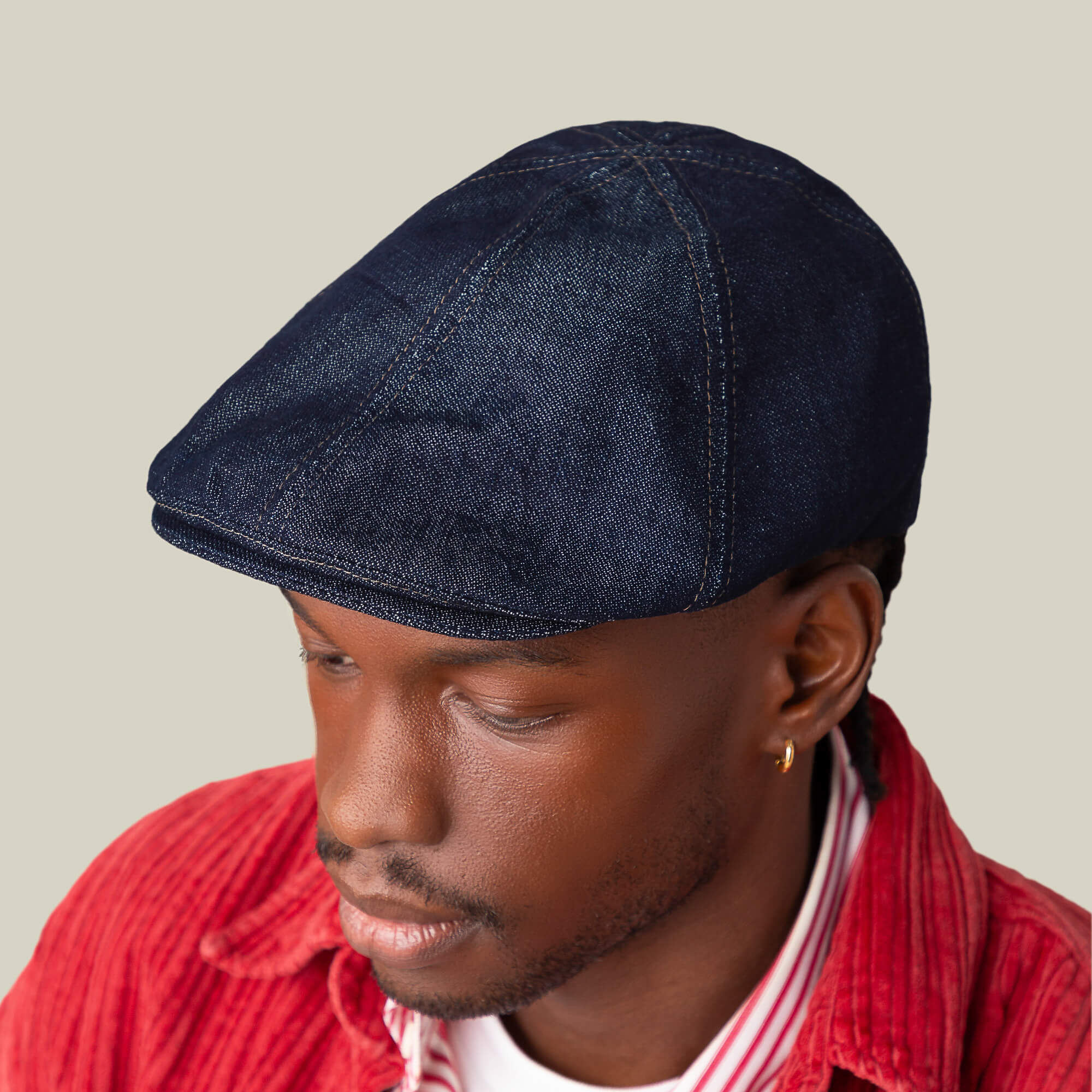 A person looks downward against a plain background, wearing Goorin Bros.' Noisy Benjamin Paul dark denim flatcap, a red corduroy shirt, and a white and red striped shirt.