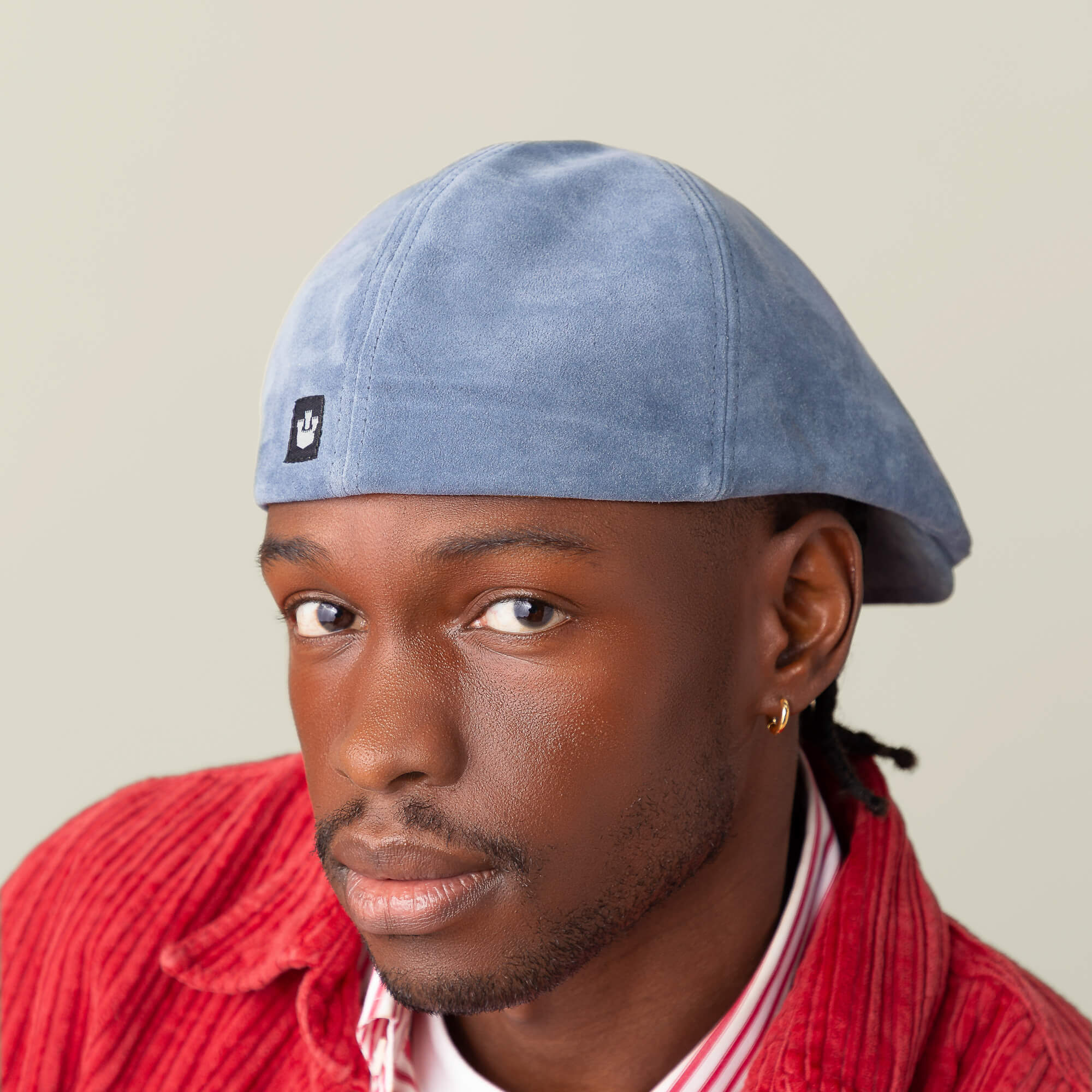 A person wearing the Cushy Benjamin Paul light blue suede flatcap by Goorin Bros. and a red corduroy jacket over a striped shirt looks toward the camera against a plain background.