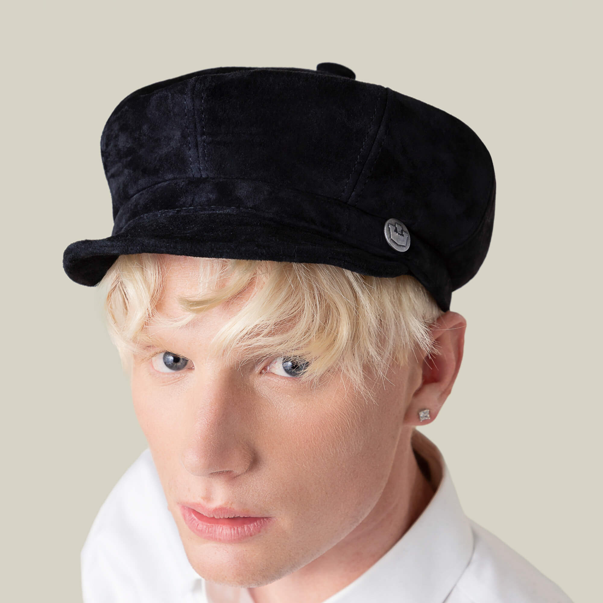 A person with blond hair wears the Ruthy black velvet cabbie flatcap by Goorin Bros., paired with a white shirt and stud earring, facing slightly to the side against a neutral background.