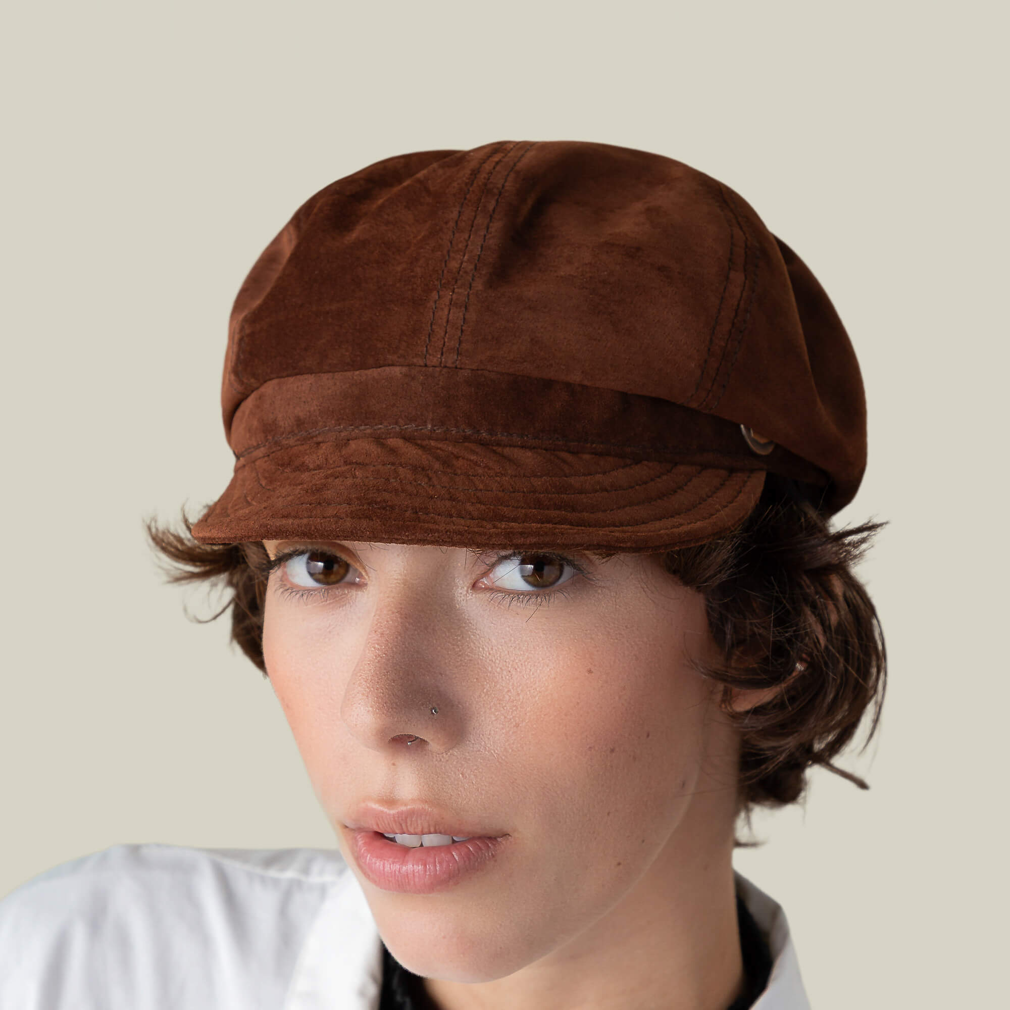 A person with short brown hair and a nose piercing wears the Ruthy suede cabbie flatcap by Goorin Bros. and a white collared shirt, posing against a plain beige background.