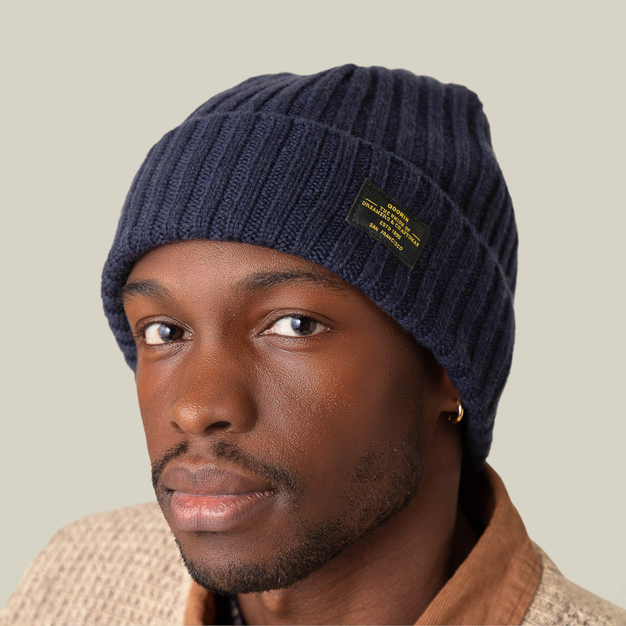 A man wearing the Goorin Bros. Dream Cap, a navy blue cashmere wool beanie with a black label, pairs it with a beige textured jacket and gold hoop earring, gazing at the camera against a plain background.