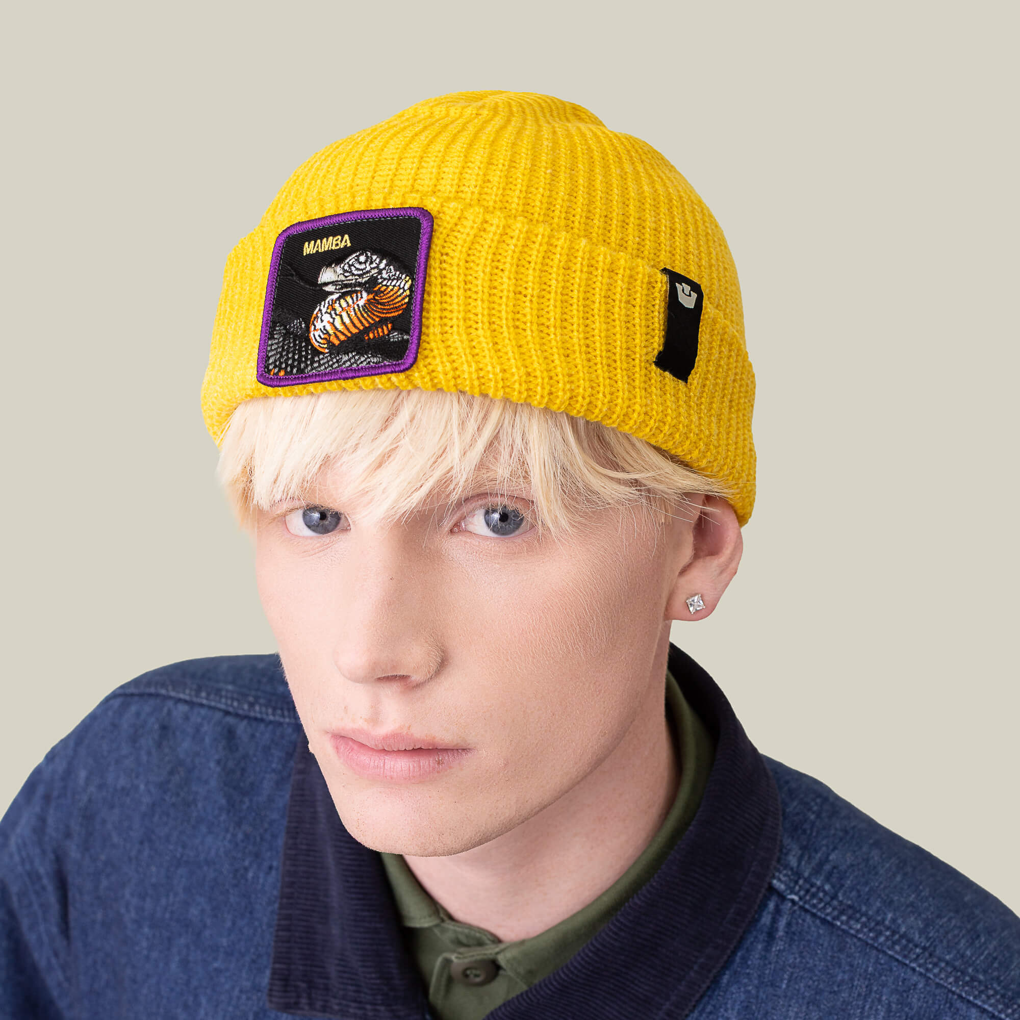 A blonde-haired individual wears The Mamba Beanie by Goorin Bros., featuring a tiger patch, styled with a dark jacket and green collared shirt against a plain background.