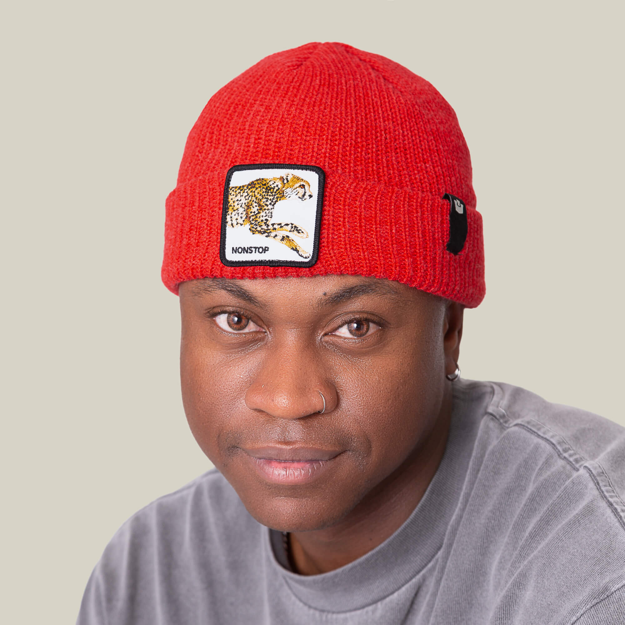 Person wearing The Nonstop Beanie by Goorin Bros.—a red knit beanie with a leopard patch—and a grey shirt, looking at the camera against a plain light background.