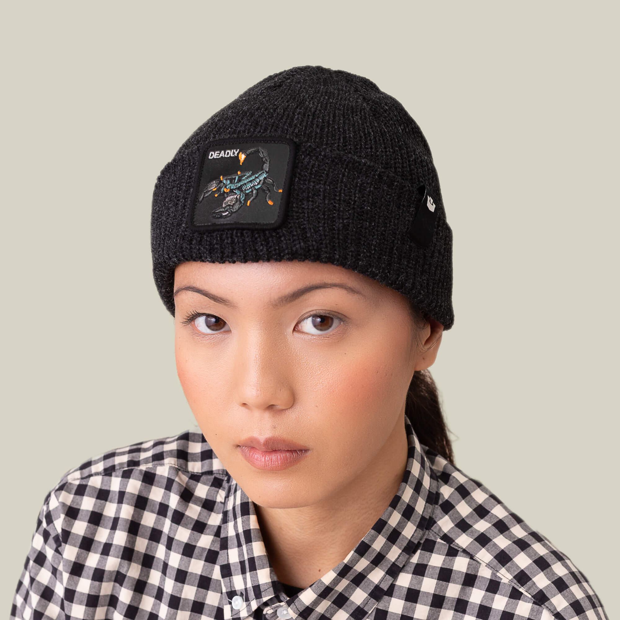 Someone wears The Deadly Beanie by Goorin Bros., featuring a scorpion patch, with a black-and-white checkered shirt, looking directly at the camera against a plain background.