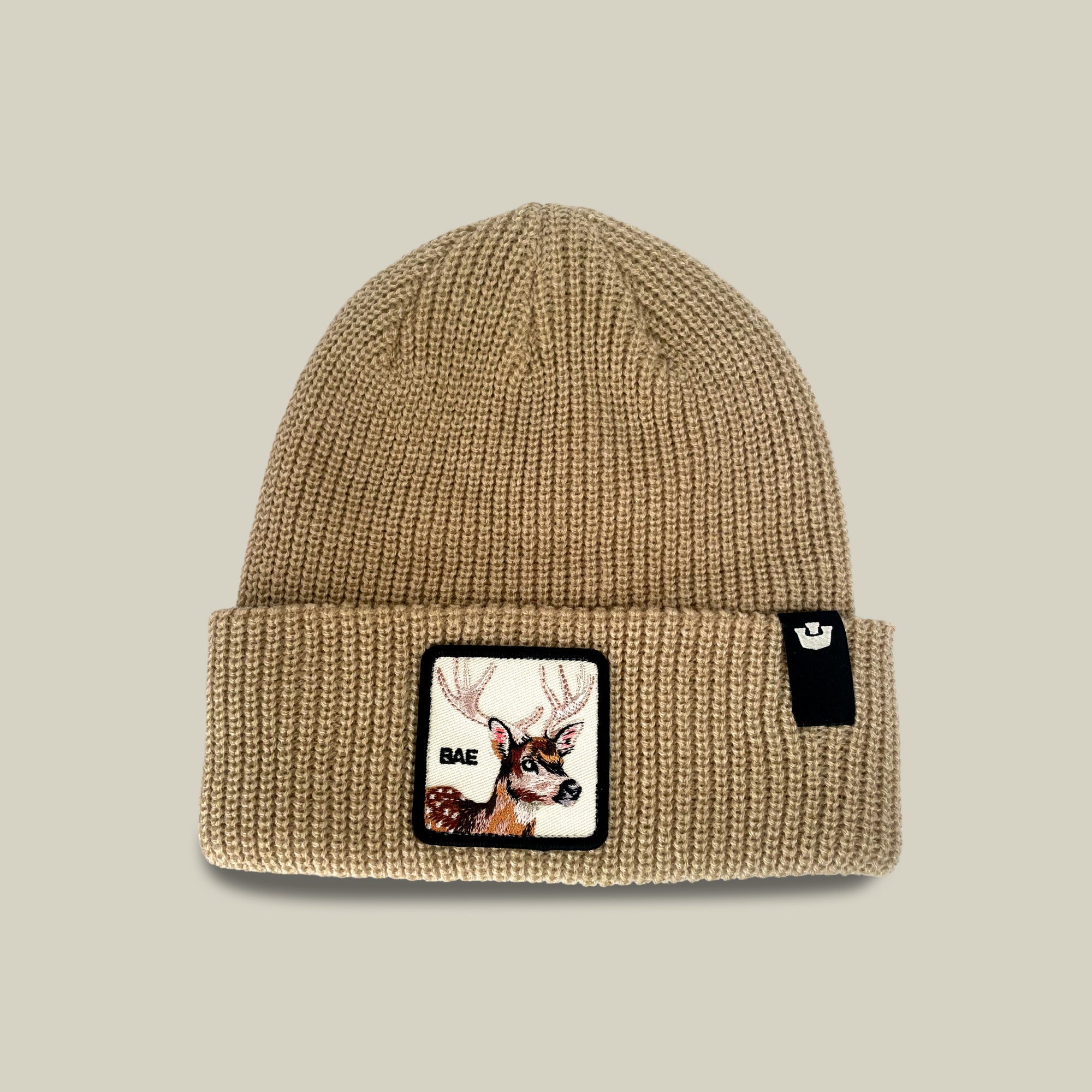 The Bae Beanie from Goorin Bros. is an everyday knit in a chalk hue, crafted from soft acrylic, and features a patch with a deer head and the word “BAE,” along with a small black tag on the cuff.