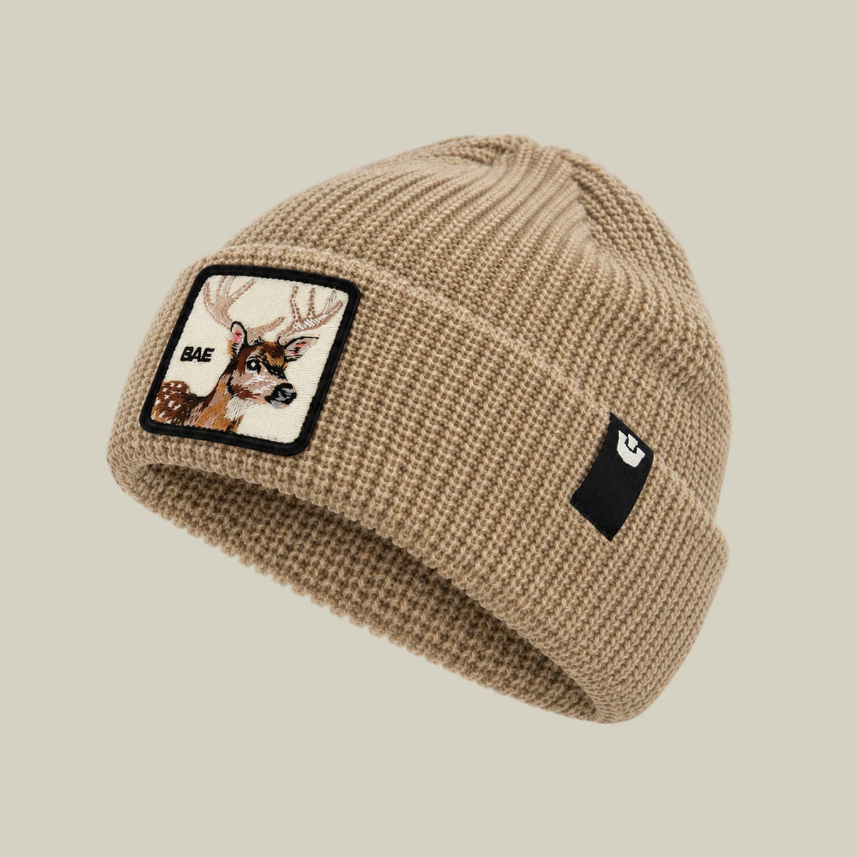 The Bae Beanie by Goorin Bros is a beige ribbed knit beanie with a deer embroidery patch on the front and a small black logo patch on the side, displayed against a neutral background.
