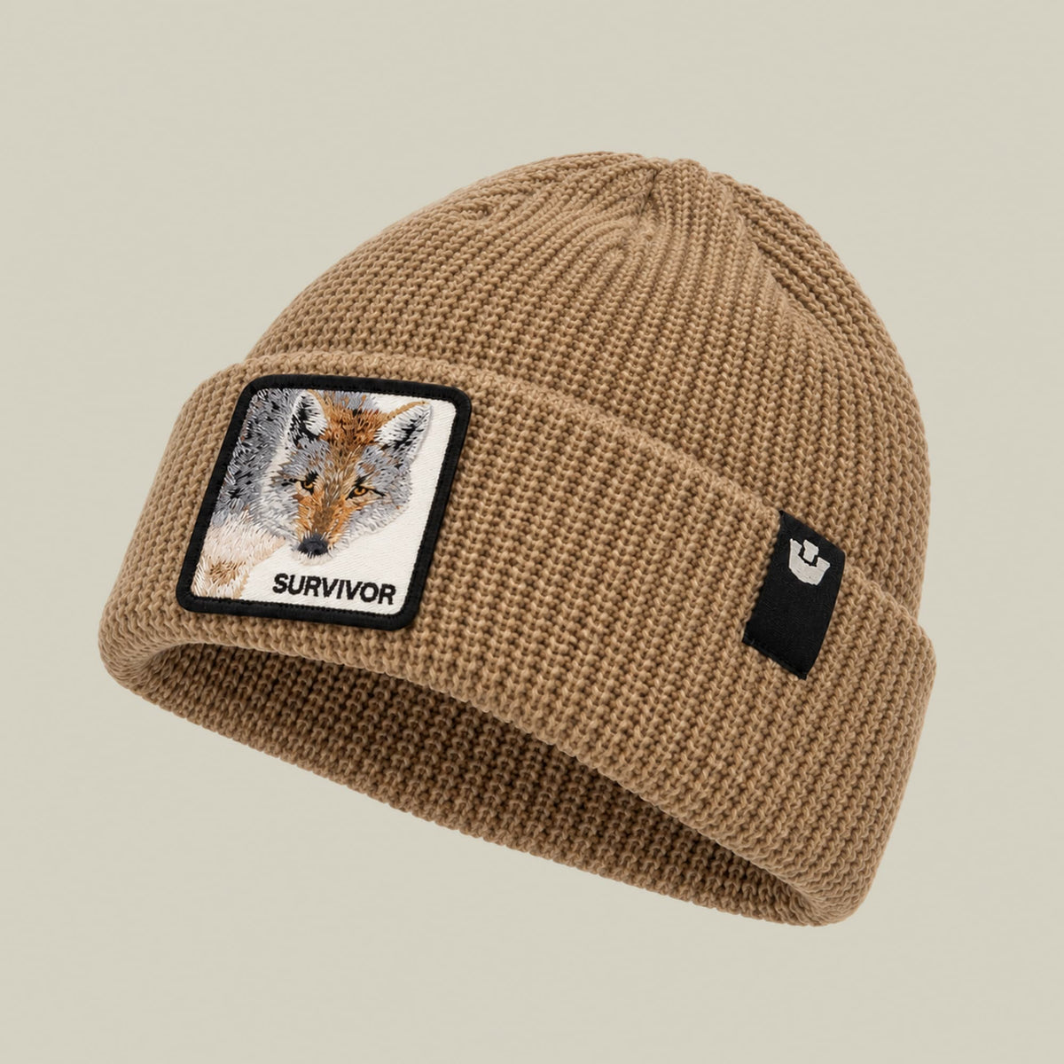 The Survivor Beanie by Goorin Bros is a beige knit beanie with a ribbed texture, featuring a embroidered fox patch with "SURVIVOR" text on the front and a small brand logo on the side, shown against a neutral background.