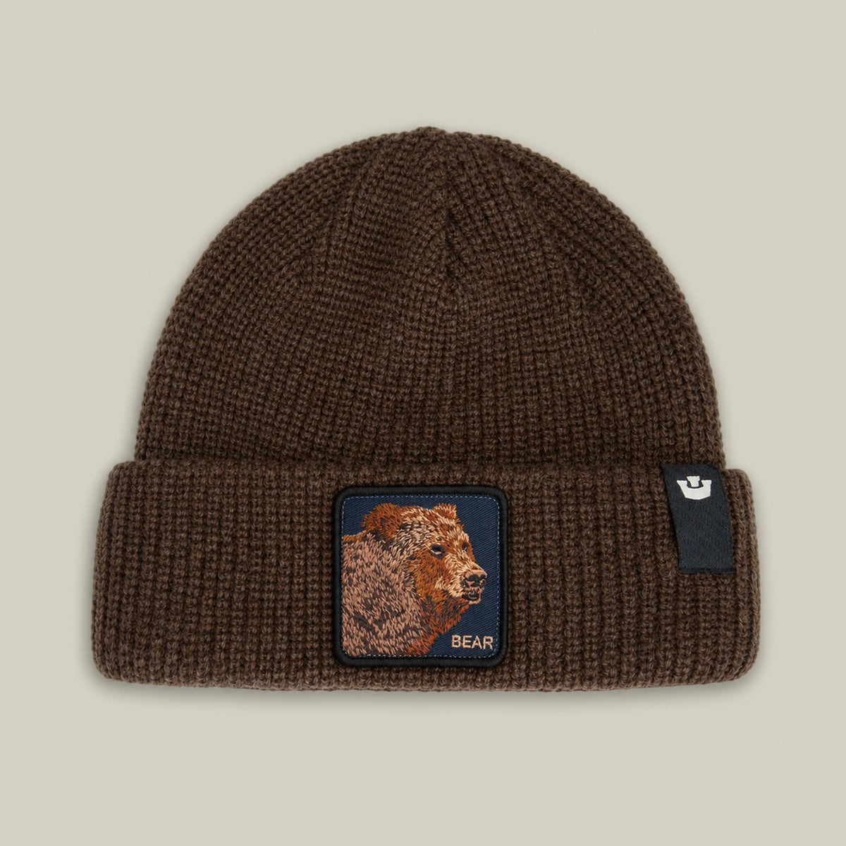 The Bear Beanie by Goorin Bros is a brown ribbed knit beanie featuring a embroidered bear patch with "BEAR" text on the front, displayed flat against a neutral background.