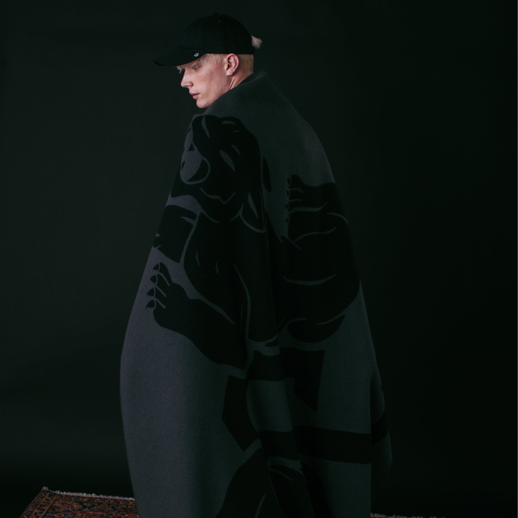 A person in a black cap stands against a dark background, wrapped in the Goorin Bros. Panther Crest Blanket featuring a bold animal graphic.