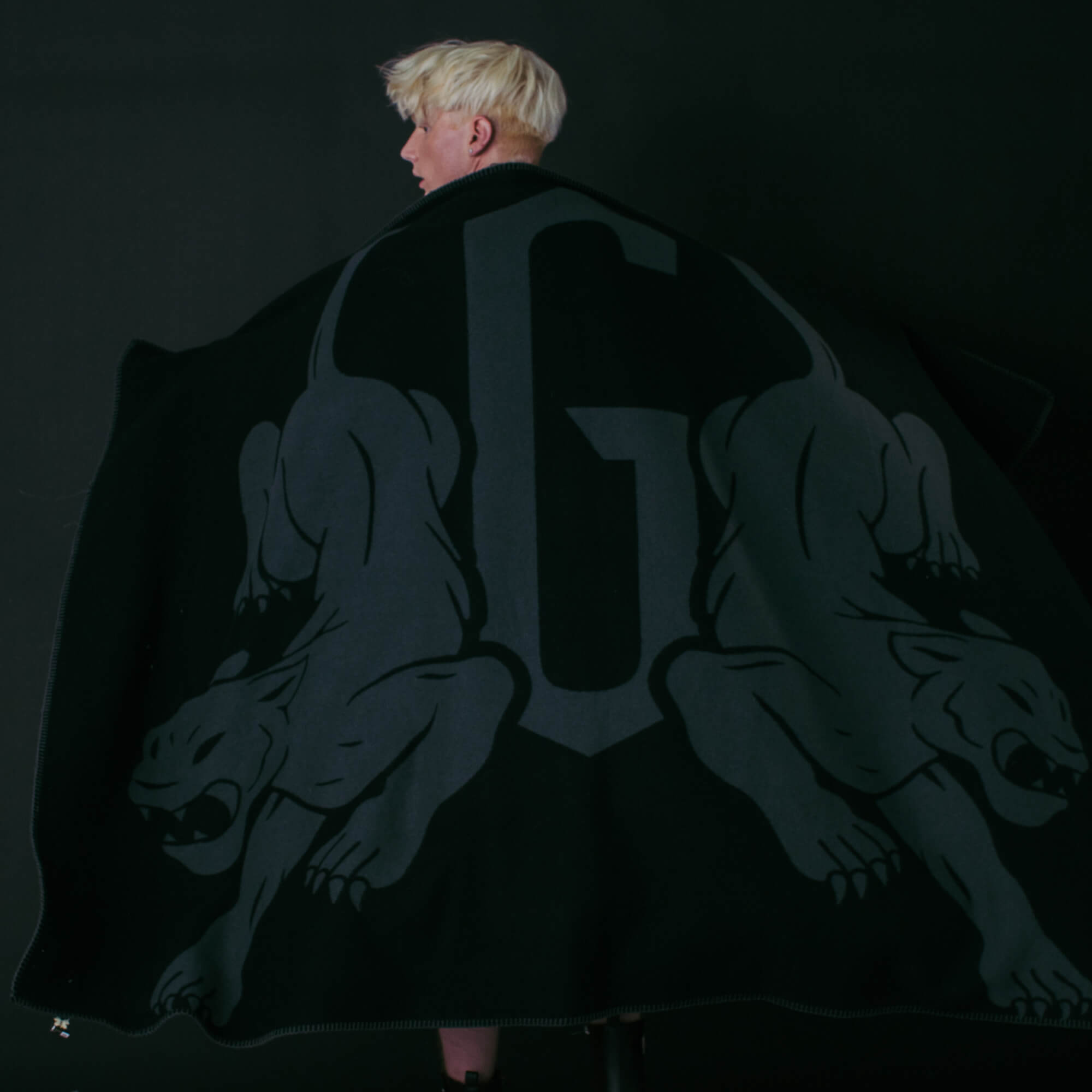 A person with short blond hair faces away from the camera, wrapped in the Panther Crest Blanket by Goorin Bros.—a black luxury throw featuring a bold 'G' and two panther-like figures on a dark background.