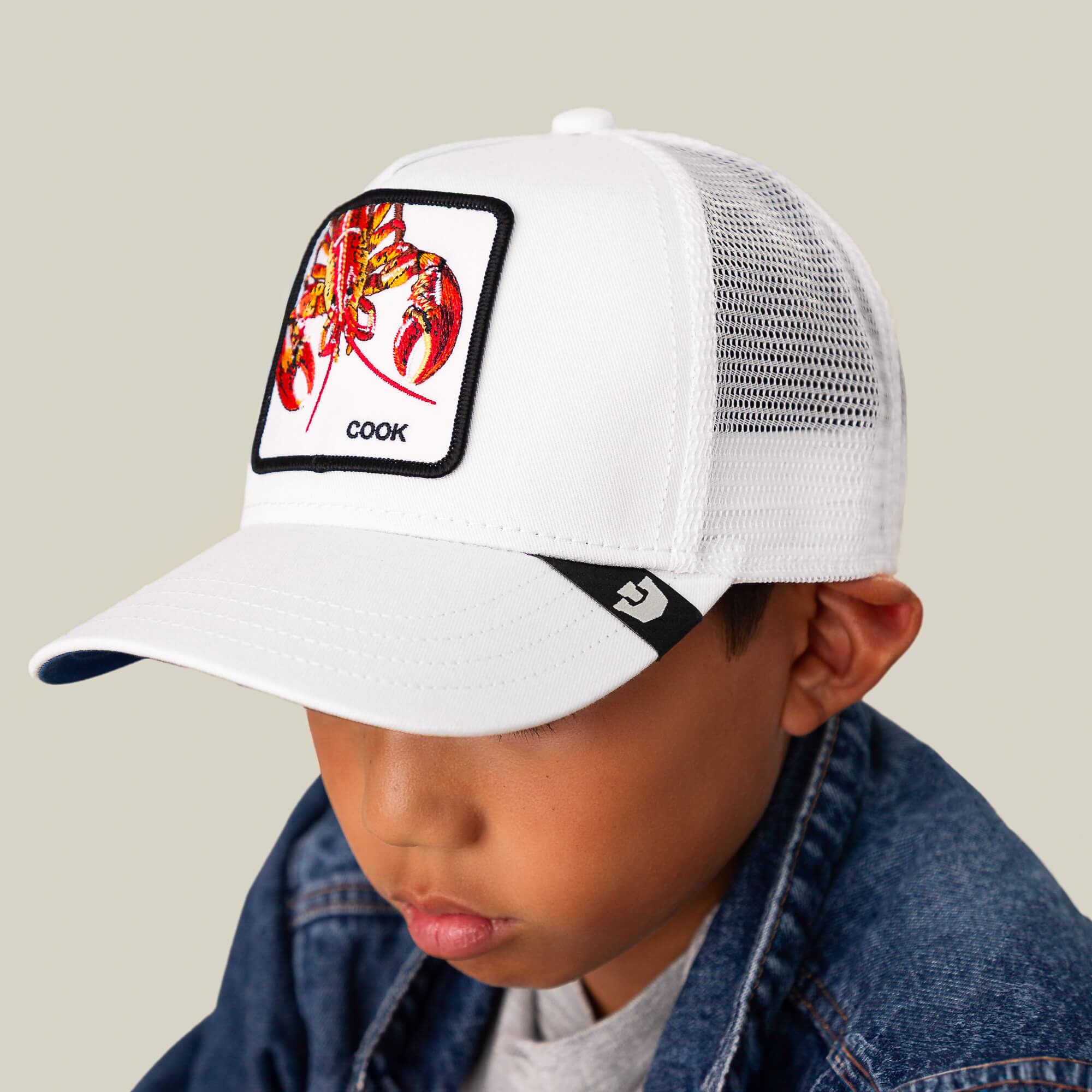 A boy wearing the Goorin Bros. Lemme Cook Mini white mesh kids hat, featuring a fiery lobster patch and "COOK," dressed in a denim jacket and gray shirt, looks downward.