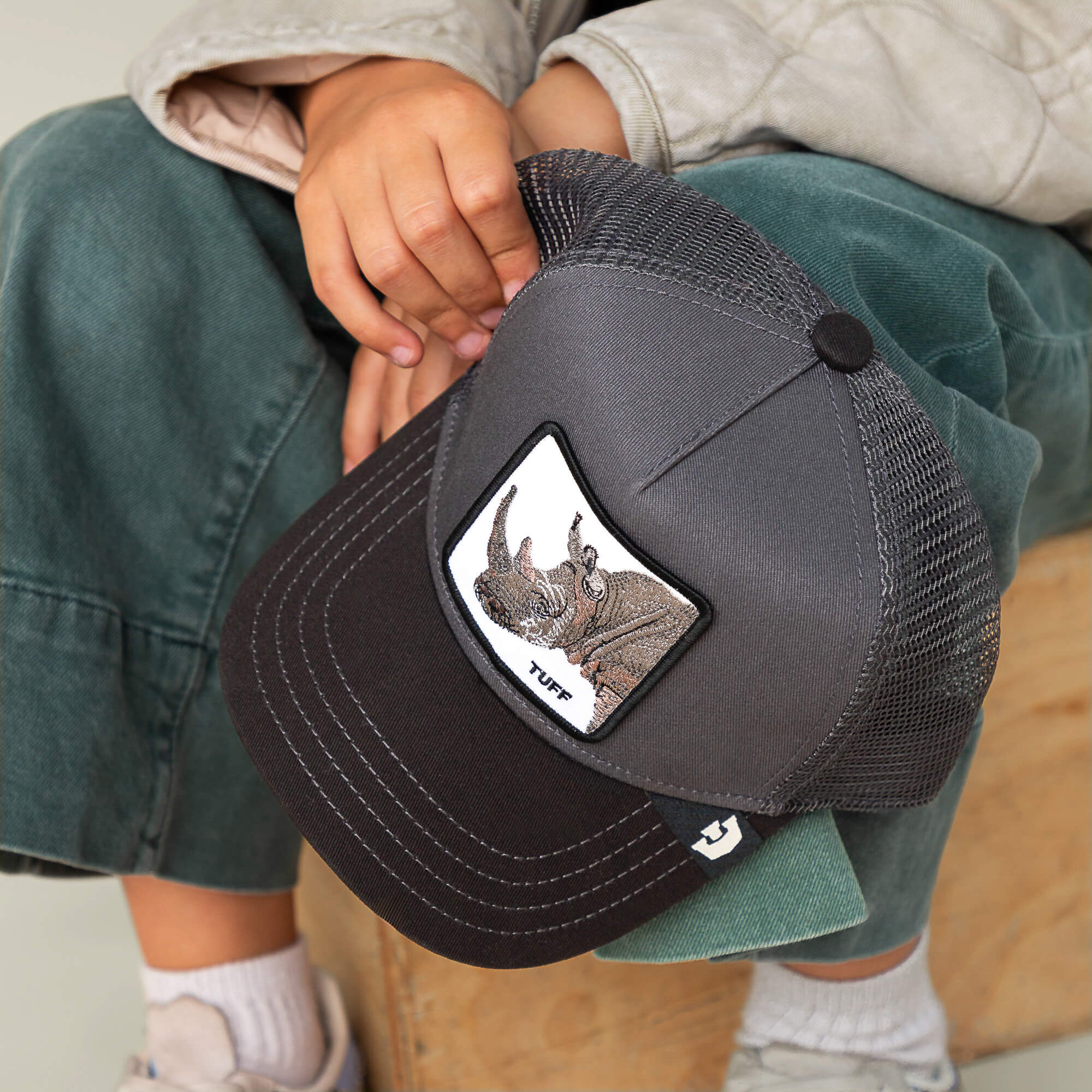 A child sits on a wooden surface holding The Tuff Rhino Mini by Goorin Bros.—a gray and black baseball cap with a rhino patch, charcoal crown, and "TUFF" on the front.