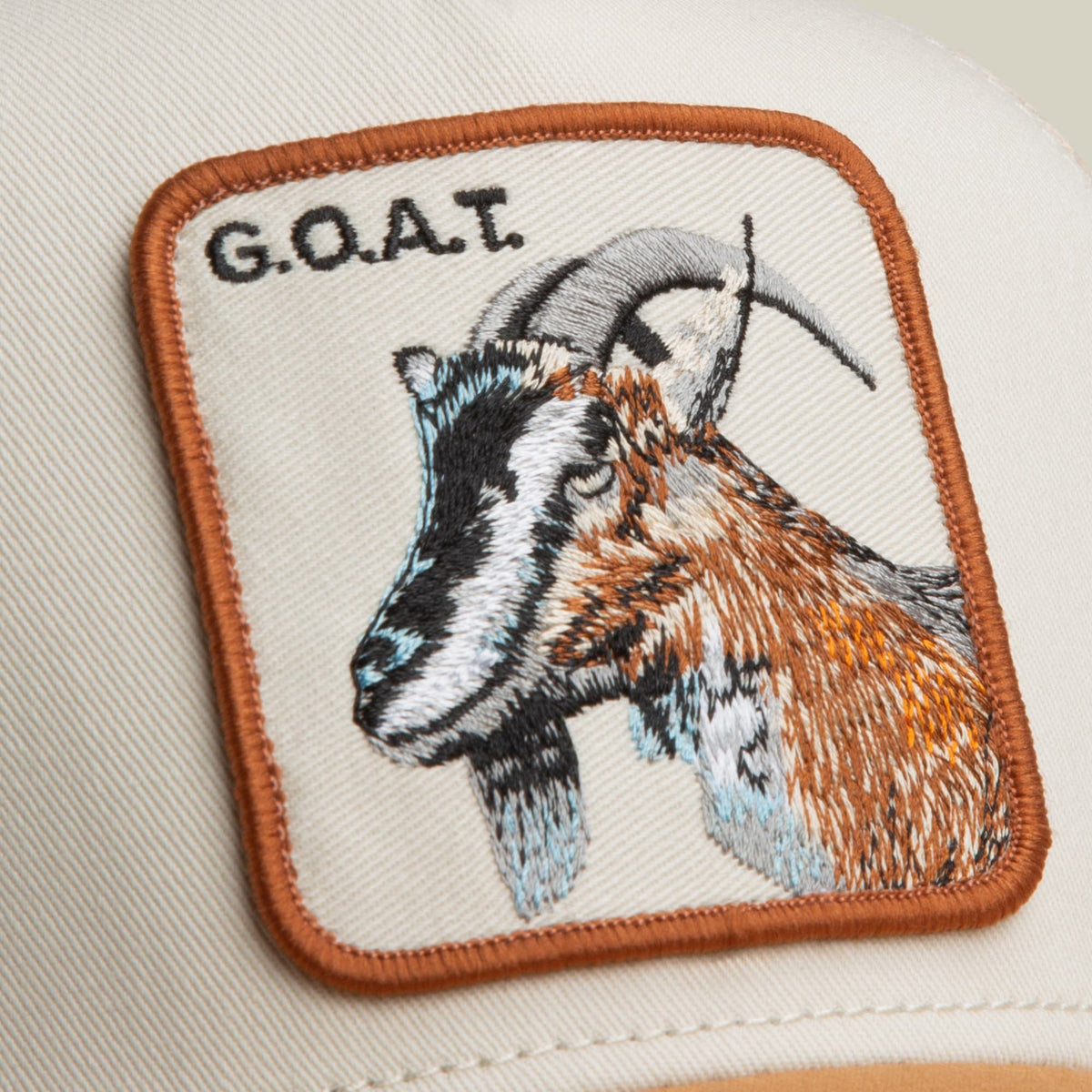 The Goorin Bros. Goat Trucker Mini is a cream-colored trucker hat with an orange-trimmed embroidered patch featuring a brown and white goat with horns in profile, shown in a flat lay studio shot.