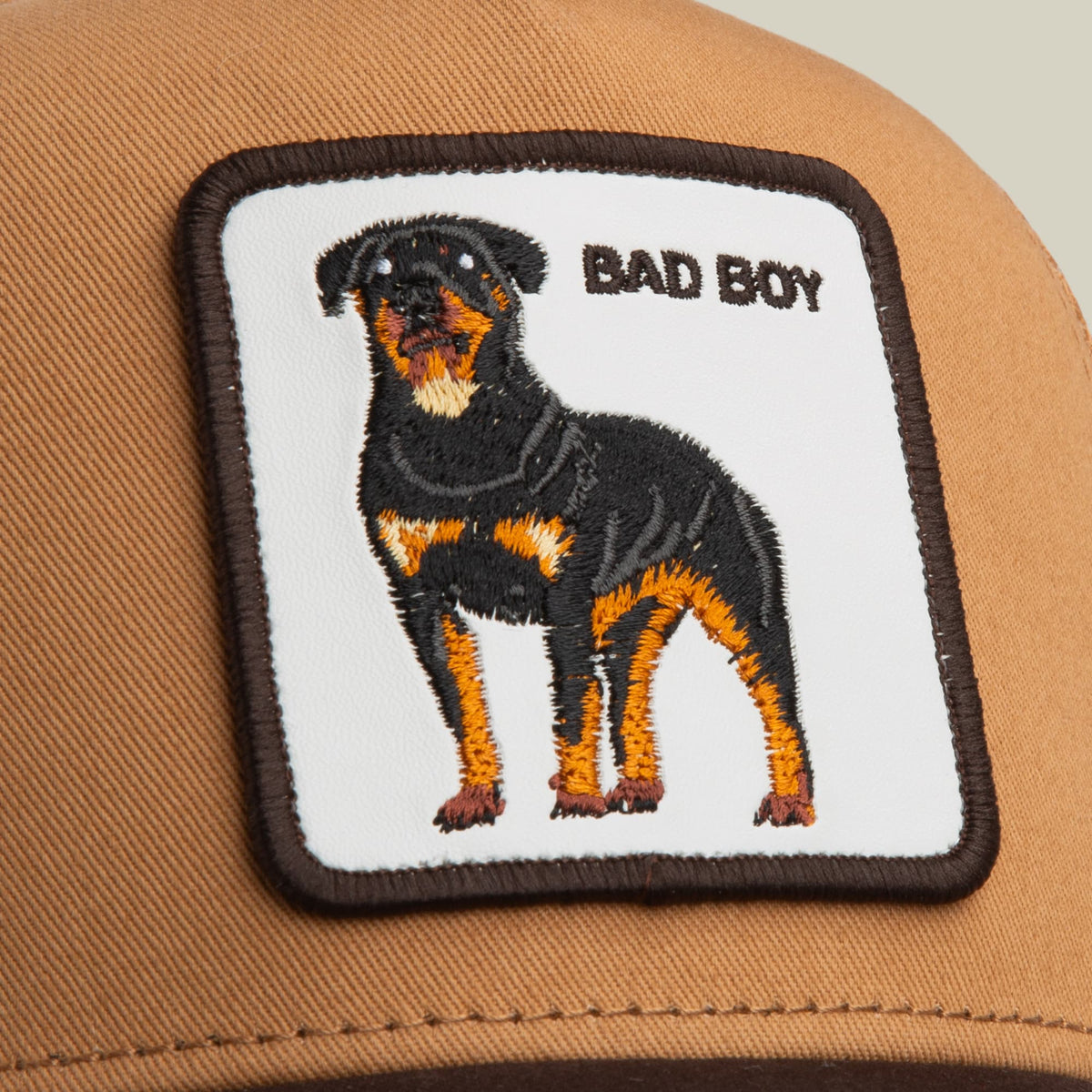 The Goorin Bros. Bad Boy Trucker Mini is a tan and brown mesh trucker cap featuring an embroidered black rottweiler with orange and tan markings and "Bad Boy" text on the front patch, shown in a studio product shot.