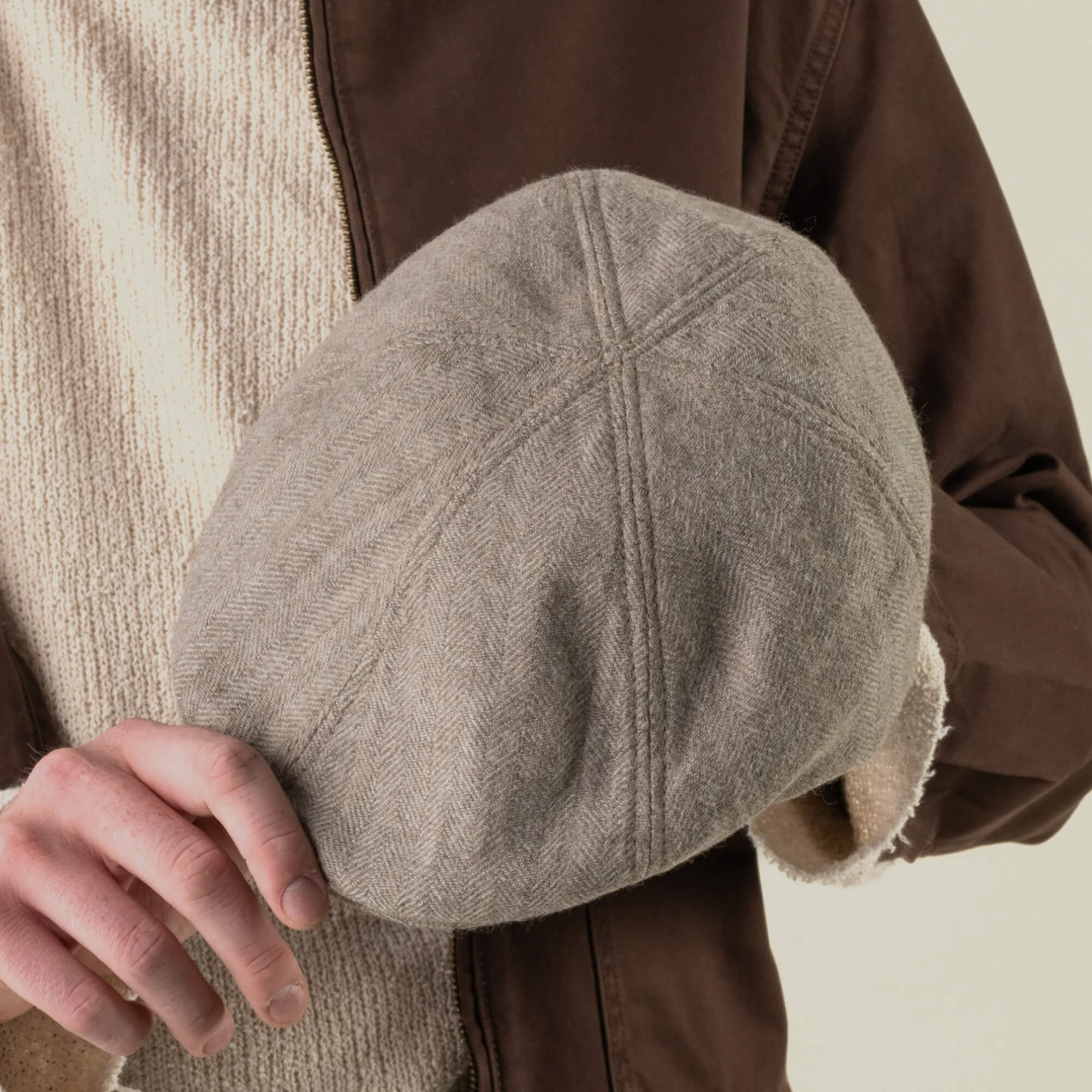 A person holds the Harry Bones flat cap by Goorin Bros. in beige herringbone, paired with a brown jacket and a textured cream sweater. This heavy-duty headwear is expertly crafted from 100% wool, providing warmth and lasting durability.