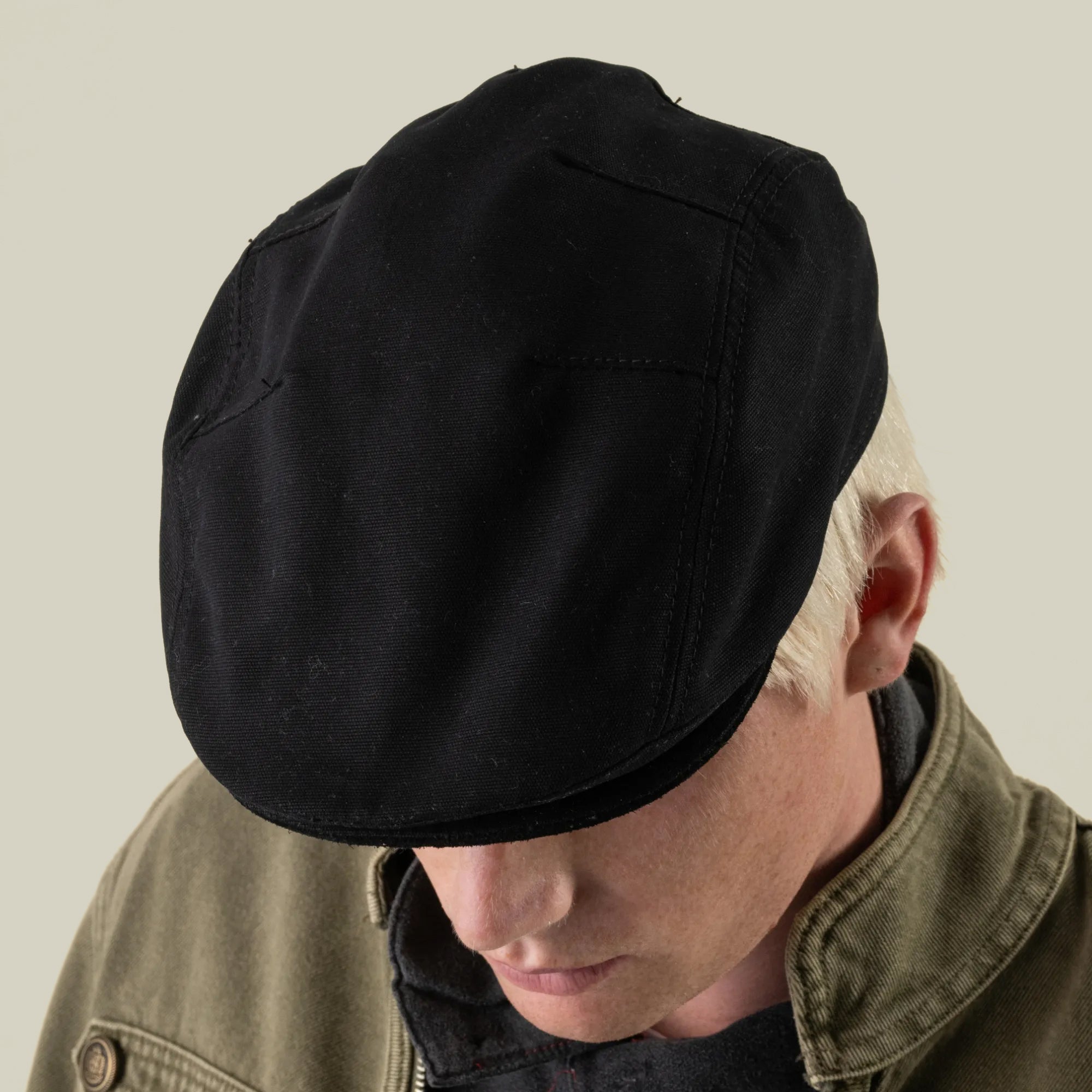 A person with light hair wears the Heatherwood black cotton flatcap by Goorin Bros., paired with an olive green jacket, looking downward against a plain background.