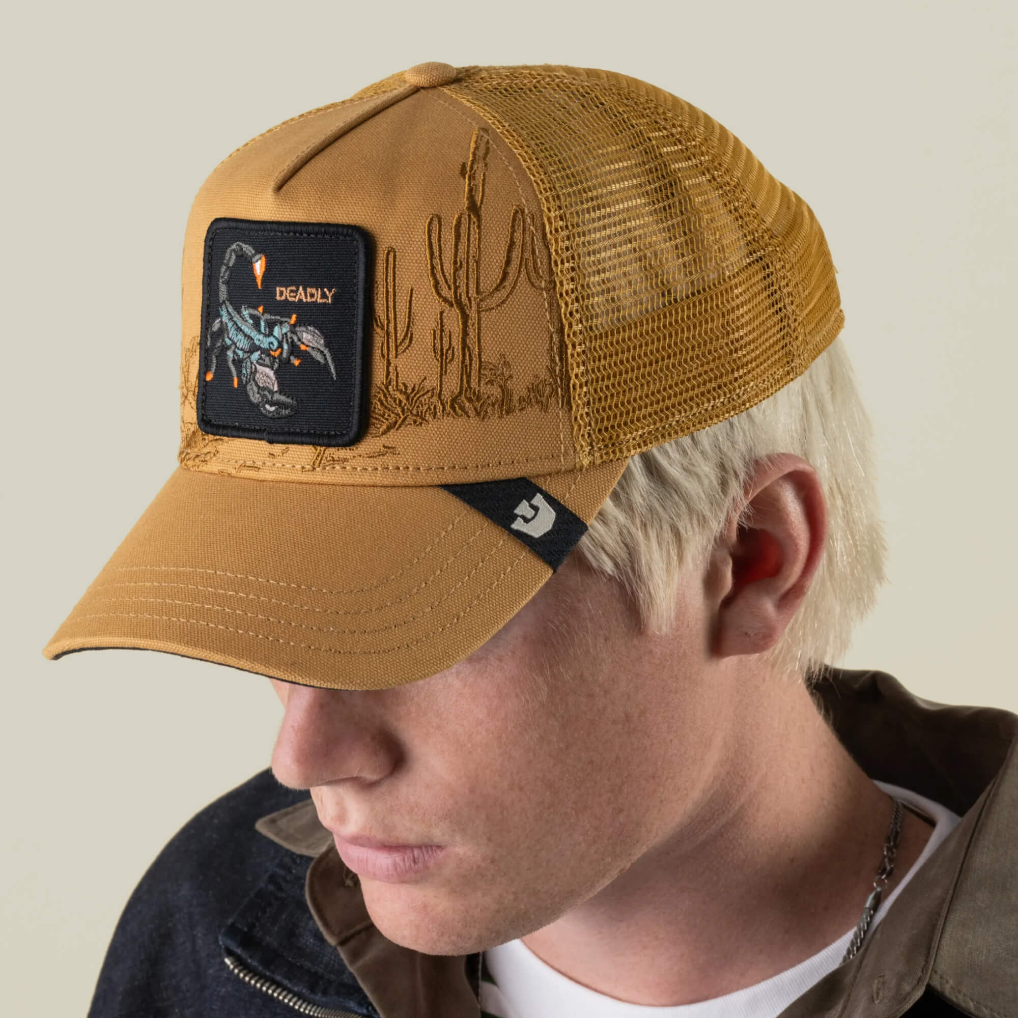 A person wearing the Goorin Bros. Badlands Scorpion tan desert trucker hat, featuring a mesh back and a distinctive cactus and scorpion patch, looks downward with their face partially obscured.