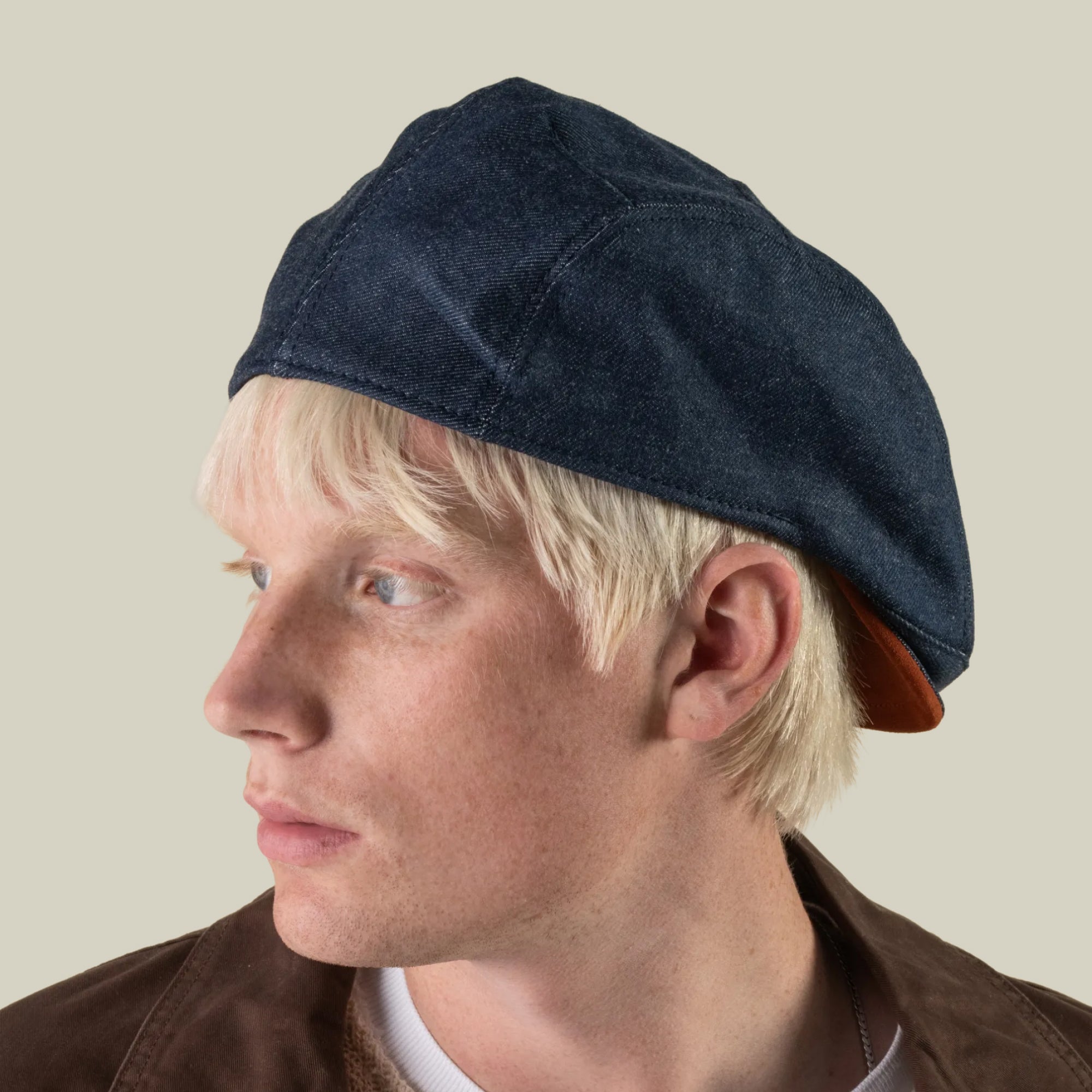 A person with light blond hair wearing the Ashfield cotton suede flat cap by Goorin Bros. and a brown jacket looks to the left against a plain light background.