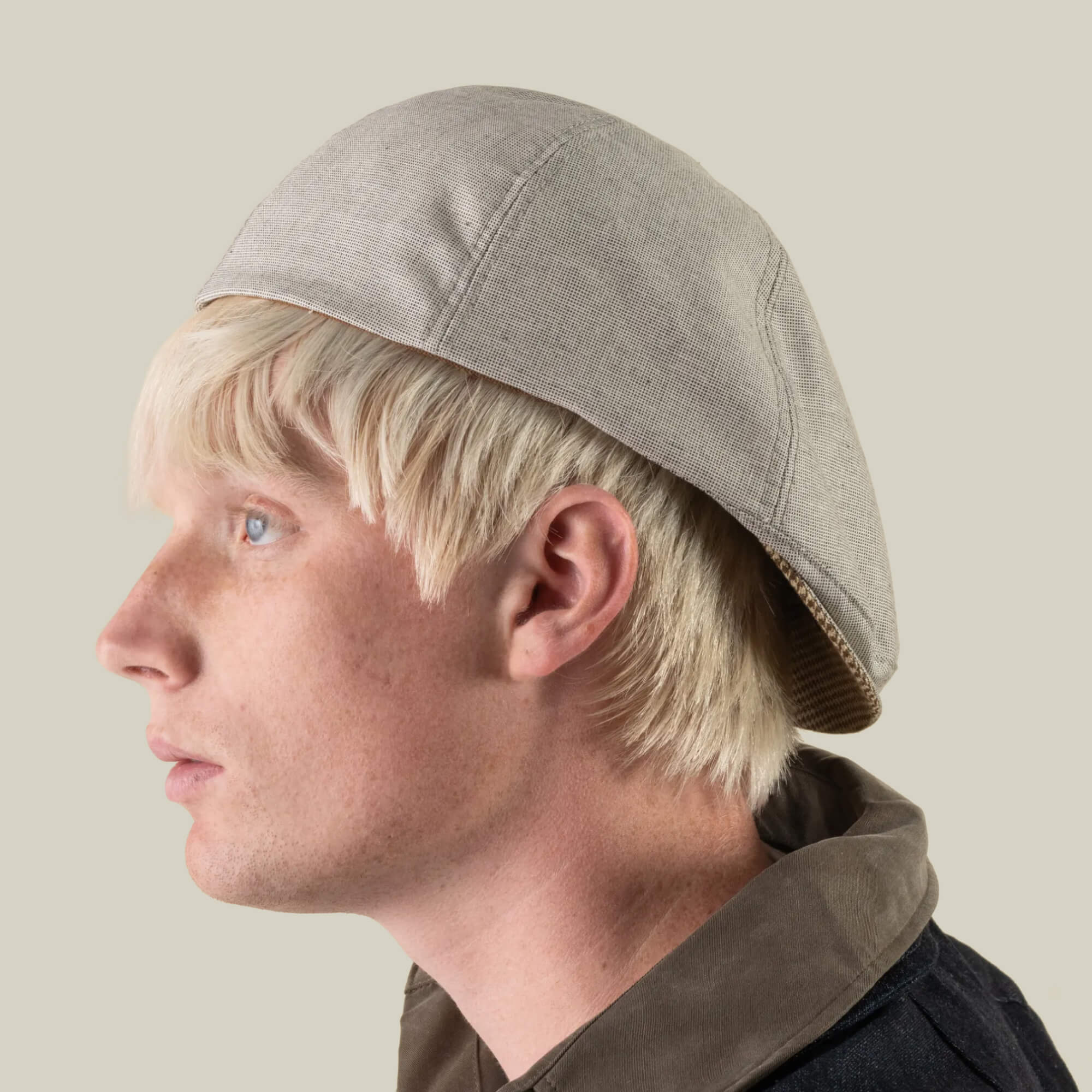 A young person with light blonde hair wears the Mile Rock 6 Panel Ivy beige cap by Goorin Bros., hand cut and sewn, styled backwards with a dark collared jacket, facing left against a plain background. This expertly crafted accessory is proudly made in the USA.
