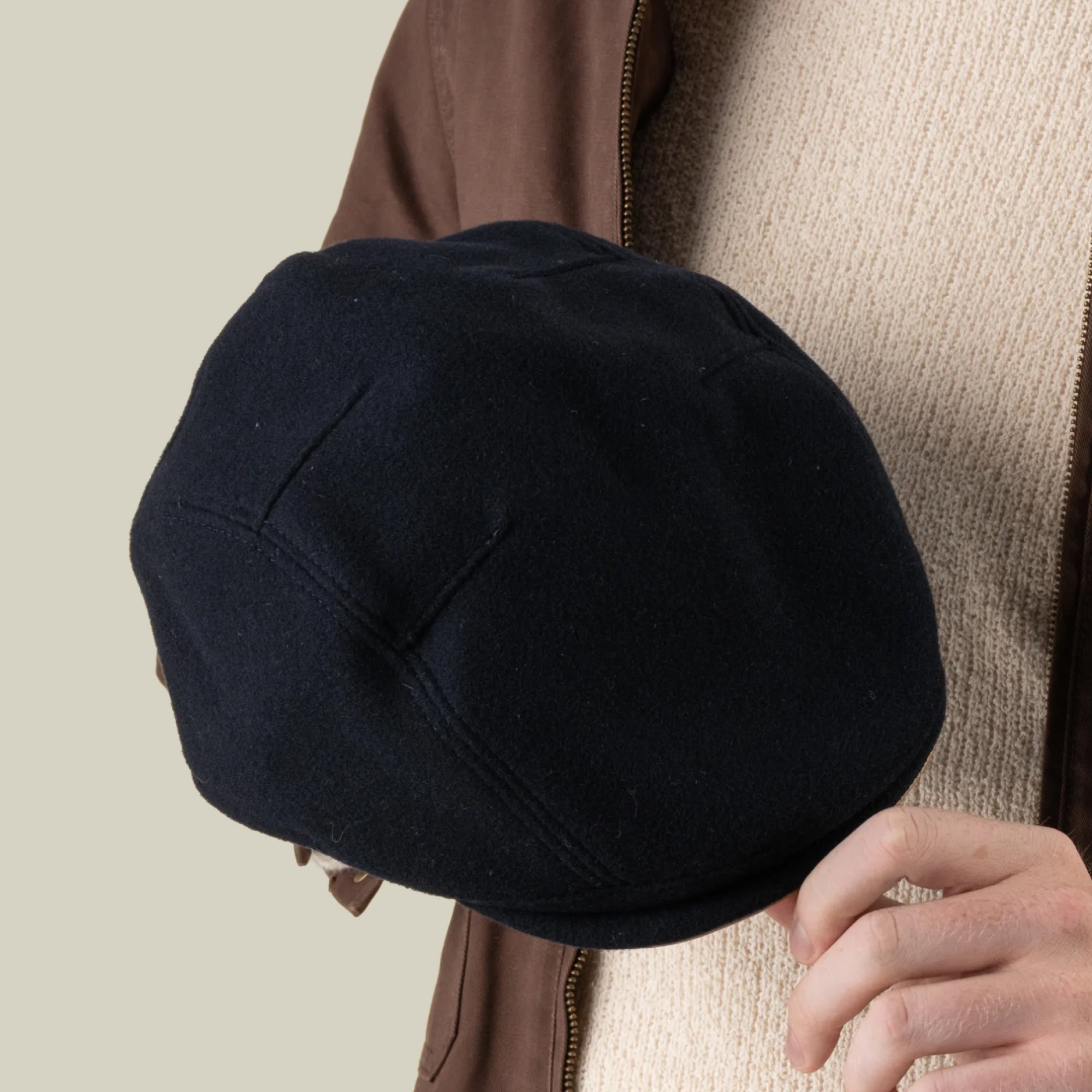 A person in a beige sweater and brown jacket holds the Goorin Bros. Cornerstone black flat cap, handmade in New Jersey, in their right hand against a plain background.