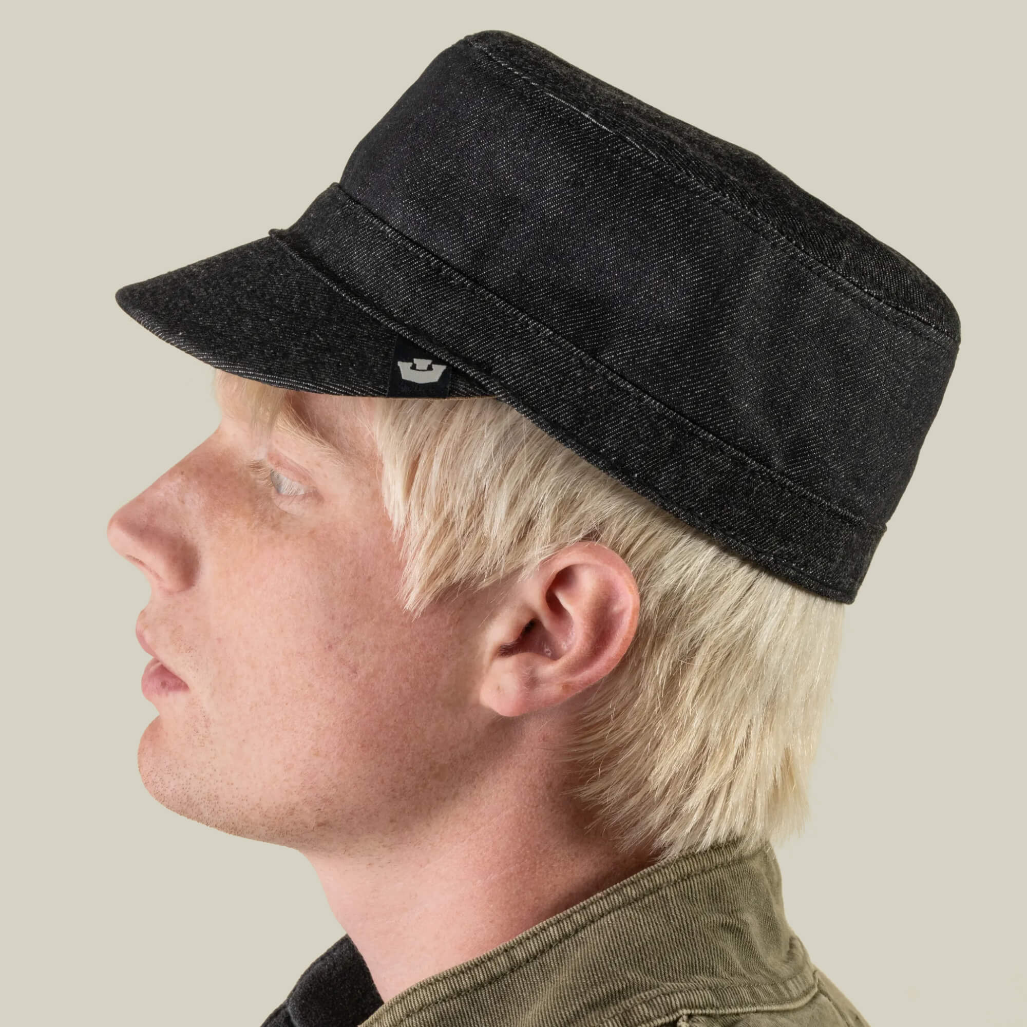 A person with light blond hair, shown in left side profile on a plain background, wears the Goorin Bros. Devonshire black cotton cadet cap—hand sewn in the USA at a New Jersey factory—paired with an olive green jacket.