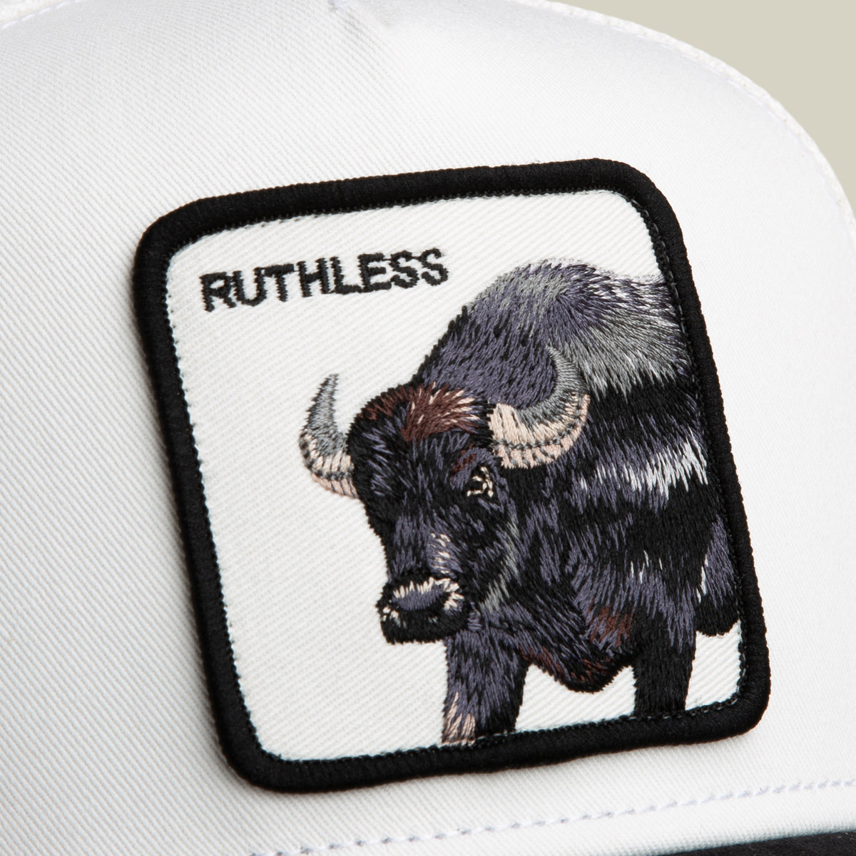 A black and white trucker cap from Goorin Bros., the Suede Ruthless features a square patch with an embroidered buffalo and the word “RUTHLESS” in black text at the top left of the patch.
