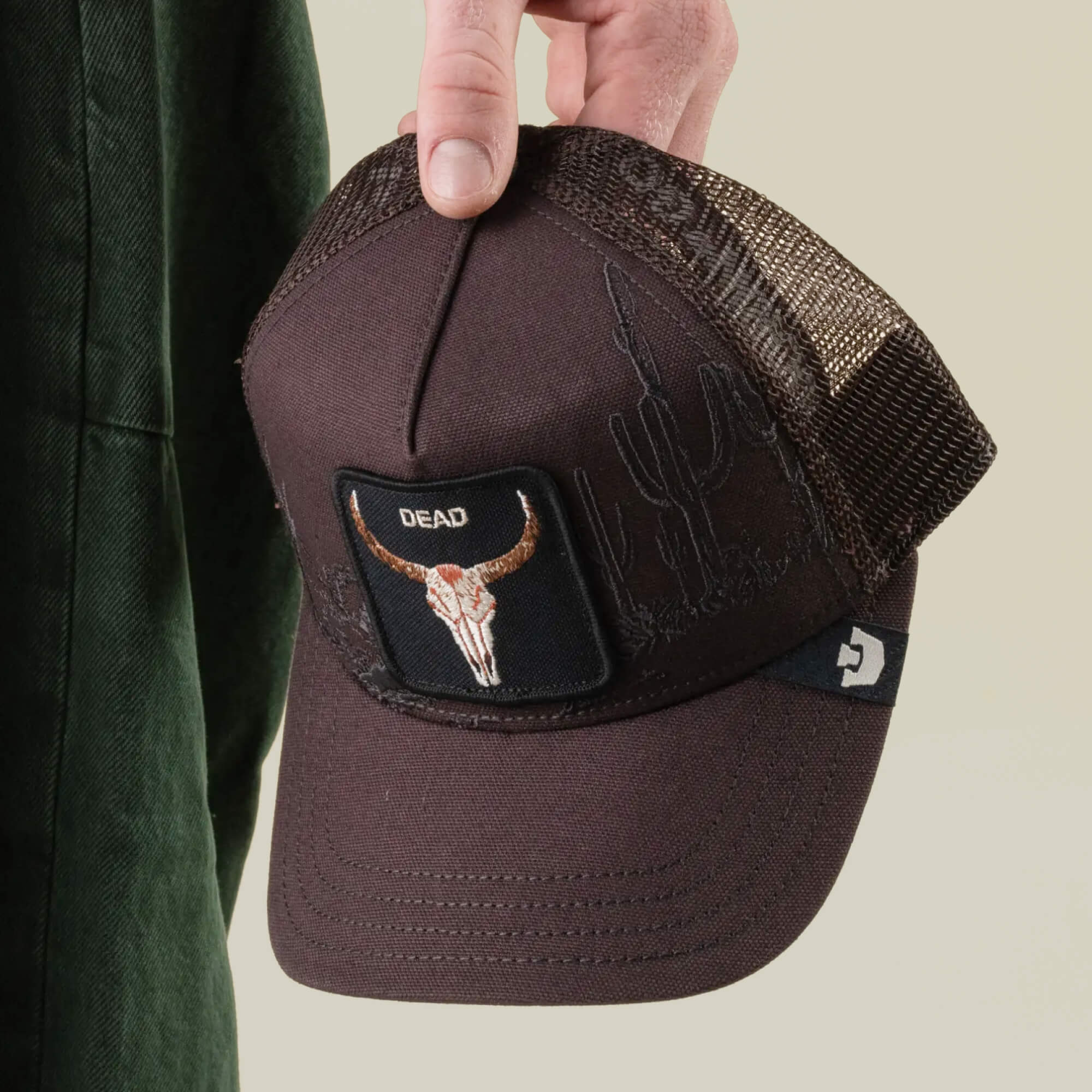 A hand holds the Goorin Bros. “Badlands Skull” hat, a brown cactus embroidery cap featuring a longhorn skull patch labeled DEAD and a breathable mesh back for comfort.