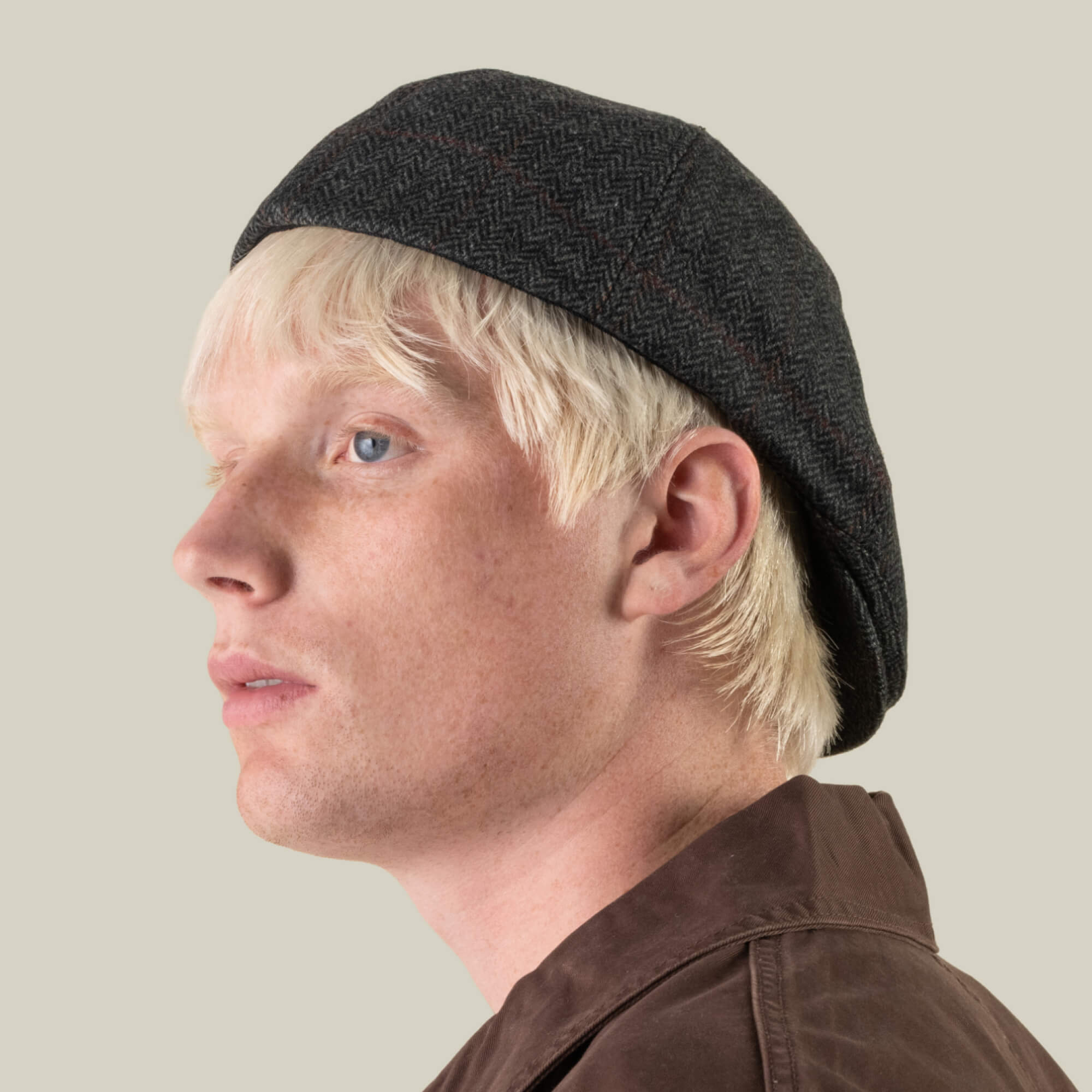 A person with pale skin and blonde hair wears the Goorin Bros. “Harry Bones” dark, textured 100% wool beret and a brown jacket, looking to the left against a neutral background.