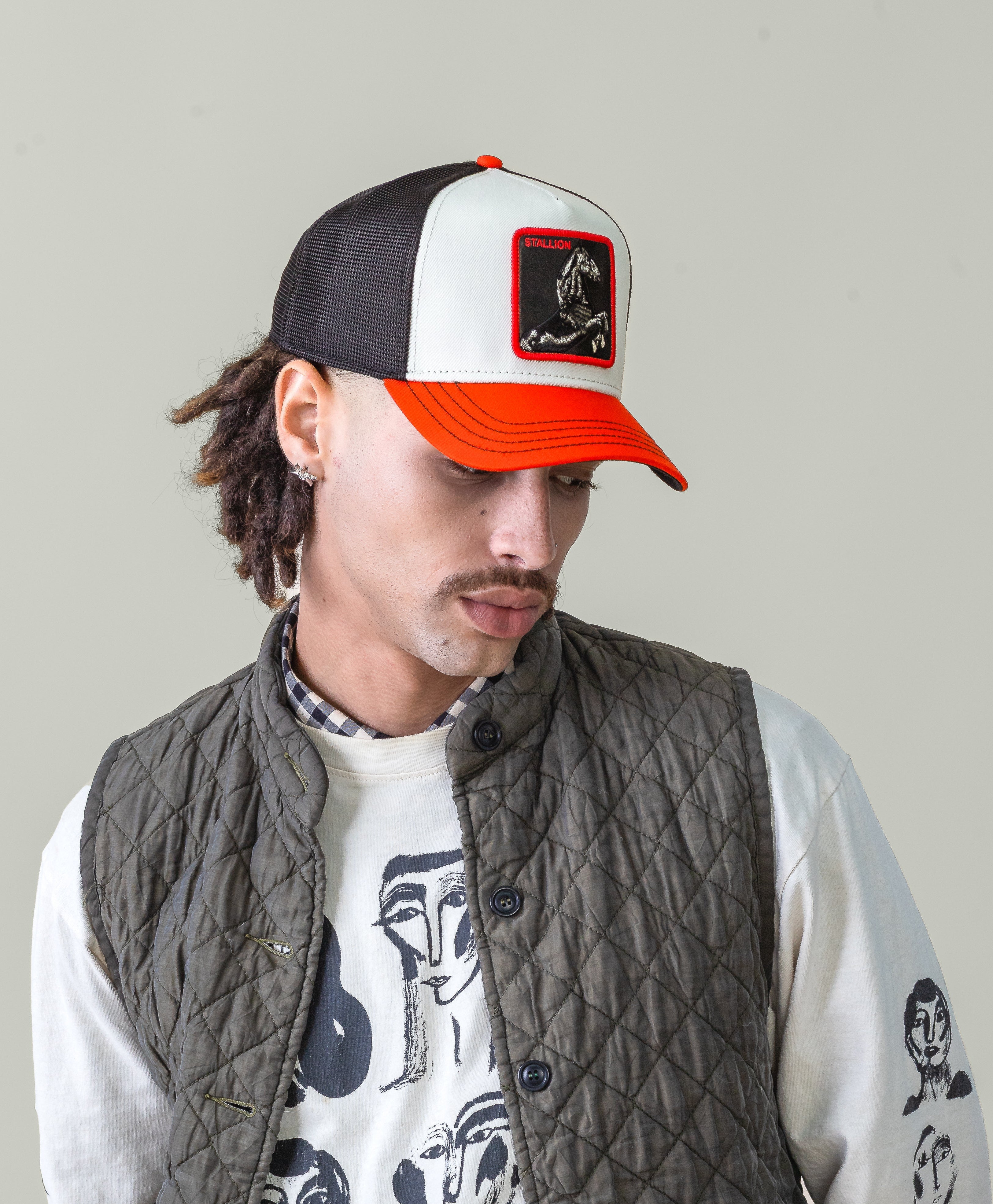 Man wearing a white graphic t-shirt, gray quilted vest, and black and orange cap on a plain background