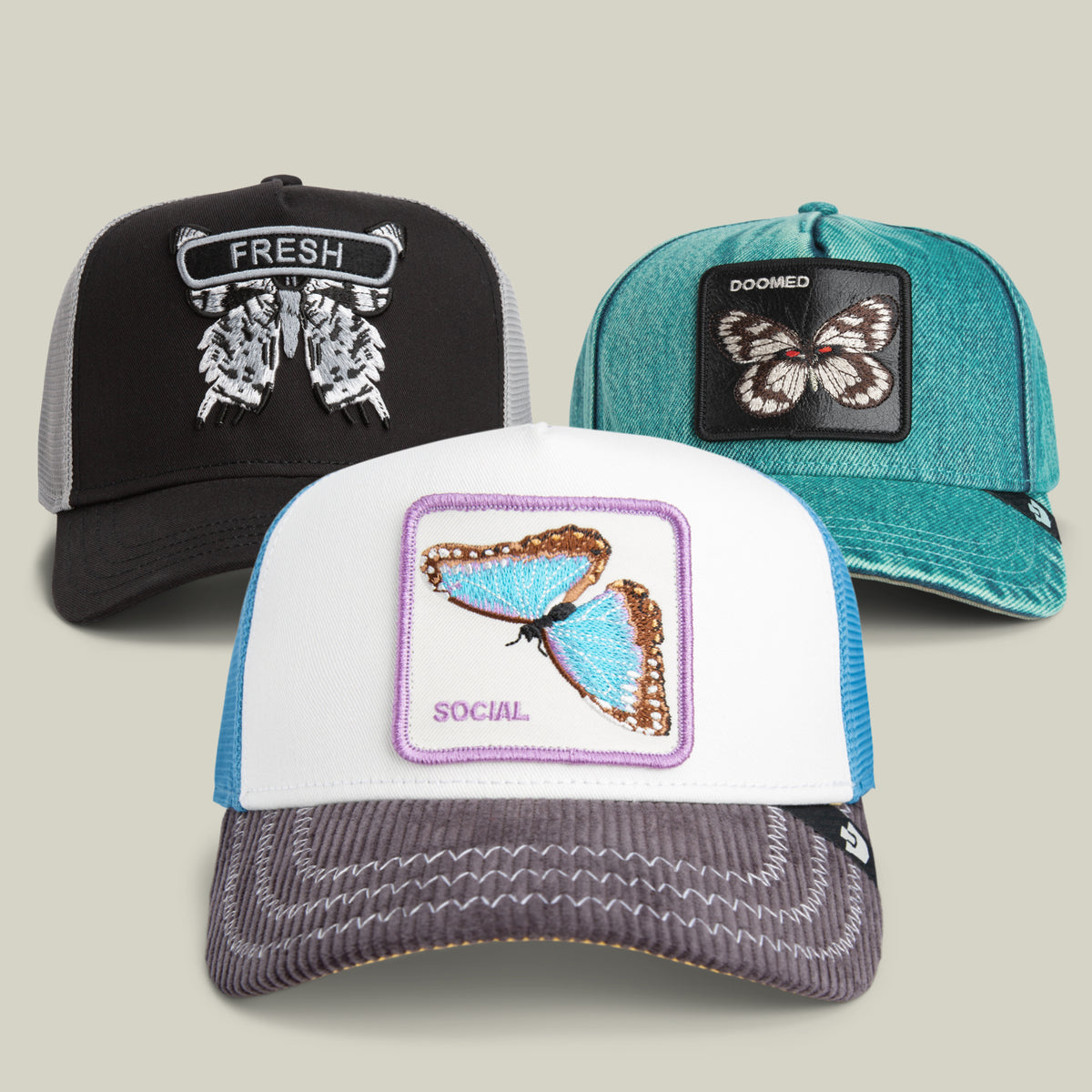 Three trucker hats from the Goorin Bros. “I Can’t Believe It’s All Butter! Bundle”—including one with an embroidered teal butterfly Doomed patch, a black hat with a bold FRESH patch, and a white-and-blue Social style—are displayed on a neutral background.