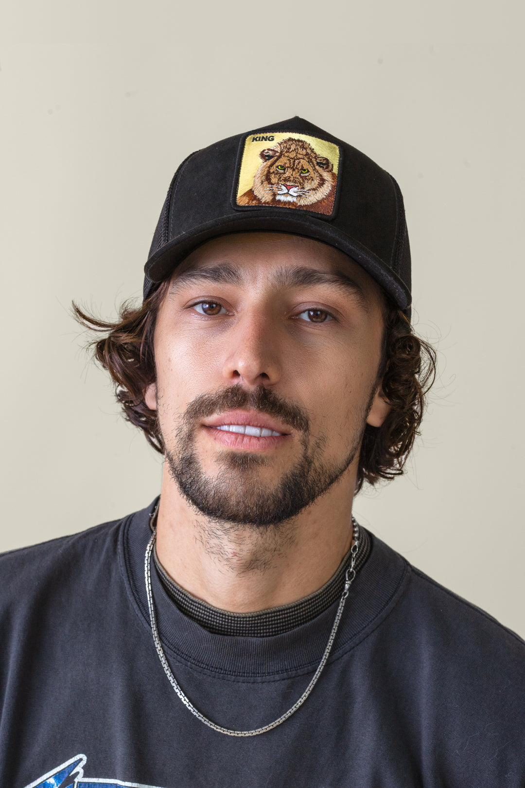 Man with medium-length brown hair and a beard wearing a black Goorin cap with a lion patch labeled King, a black shirt, and a silver chain necklace, against a plain background—showcasing the appeal of durable hats.