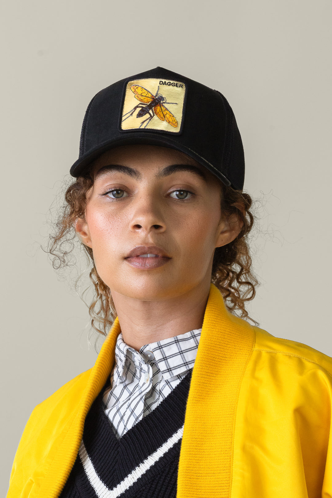A person wearing a yellow jacket, checkered shirt, and a black Goorin trucker hat with a yellow insect patch, standing against a plain background.