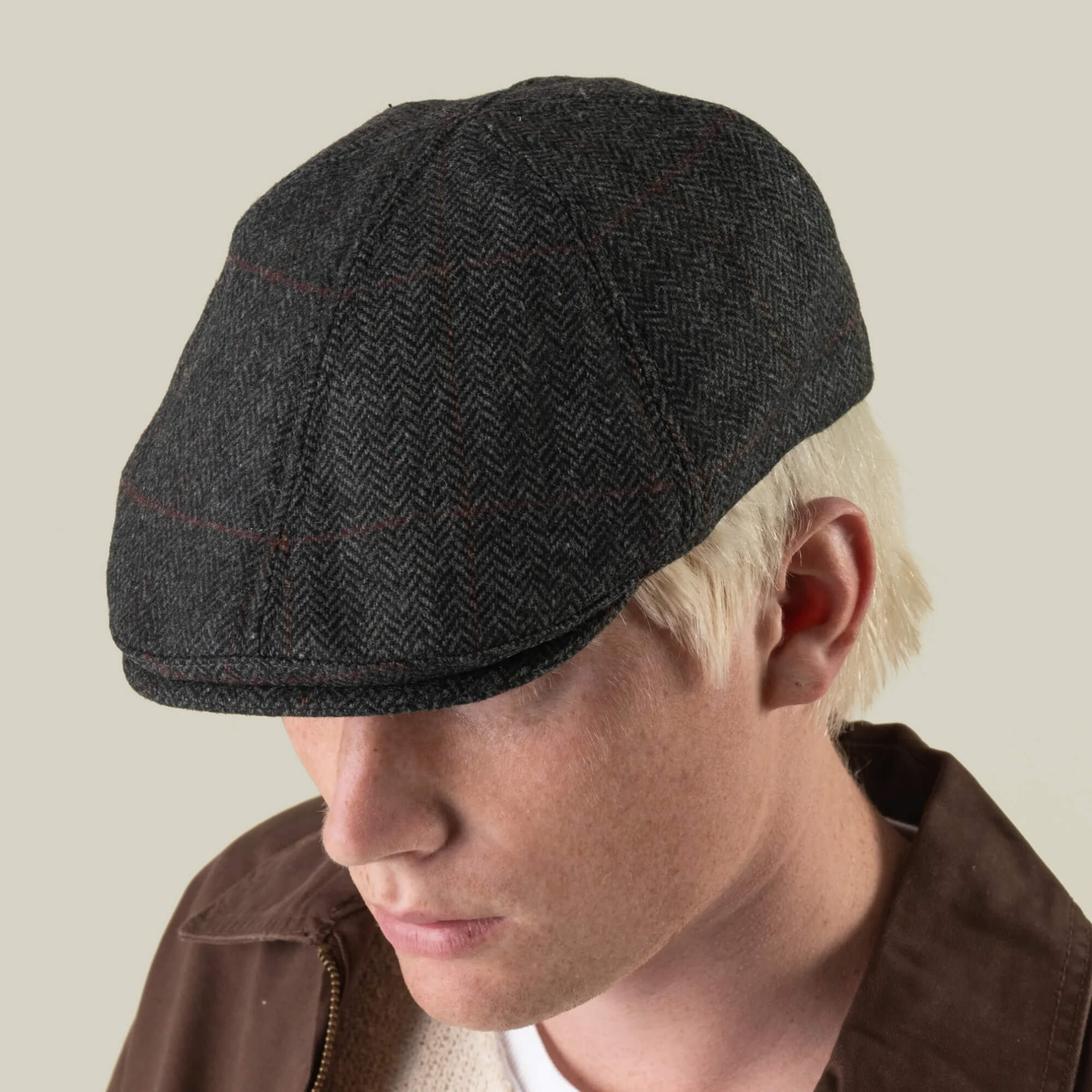 A person with light blonde hair is shown against a neutral background, wearing the Goorin Bros. Harry Bones flat cap in dark gray herringbone, expertly crafted in the USA from 100% wool. Paired with a brown jacket, this heavy-duty hat adds a classic touch to their look.
