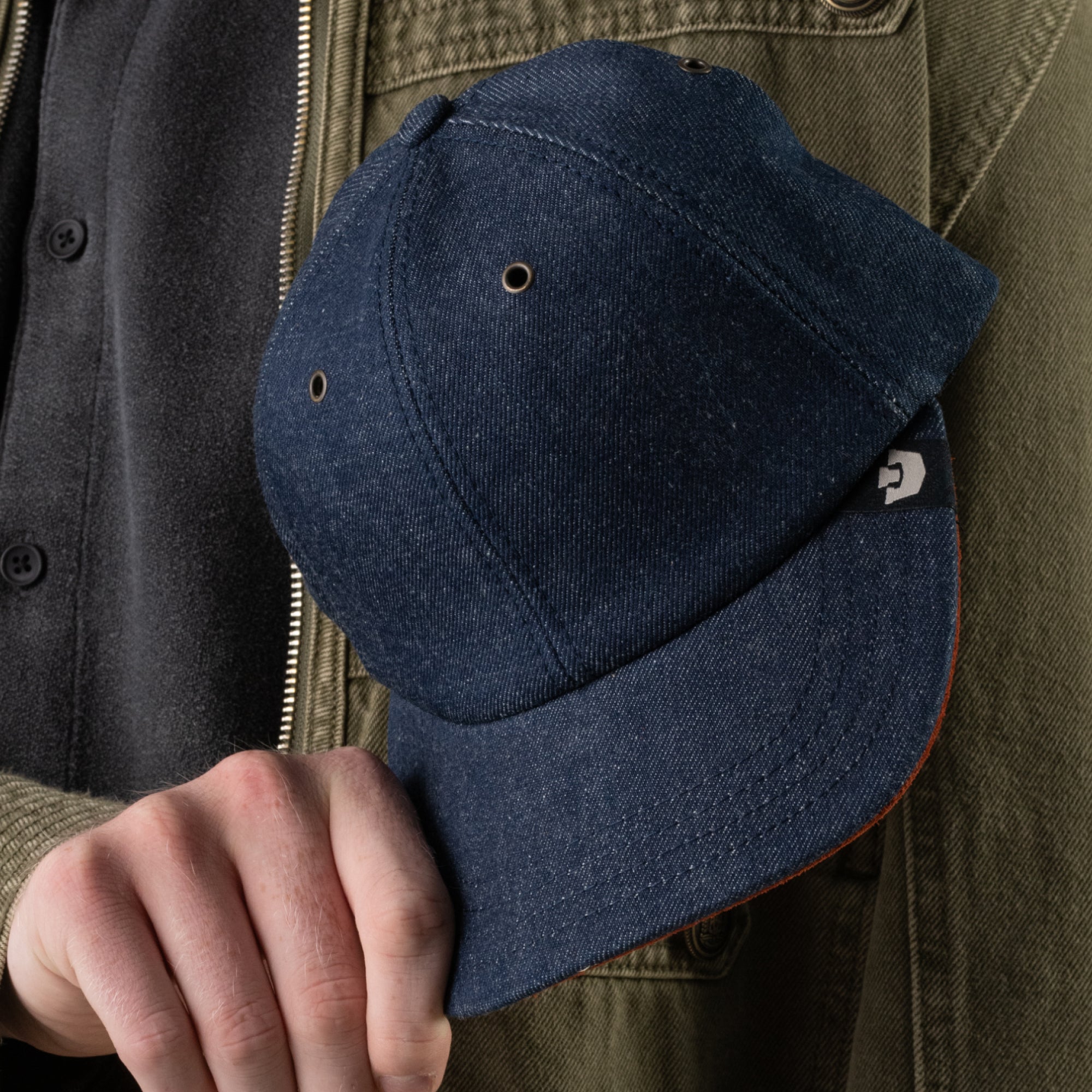 A person wearing a black shirt and olive jacket holds the Legend Ridge baseball cap by Goorin Bros.—a dark blue, mixed-material hat featuring a small tag near the brim, proudly made in the USA.