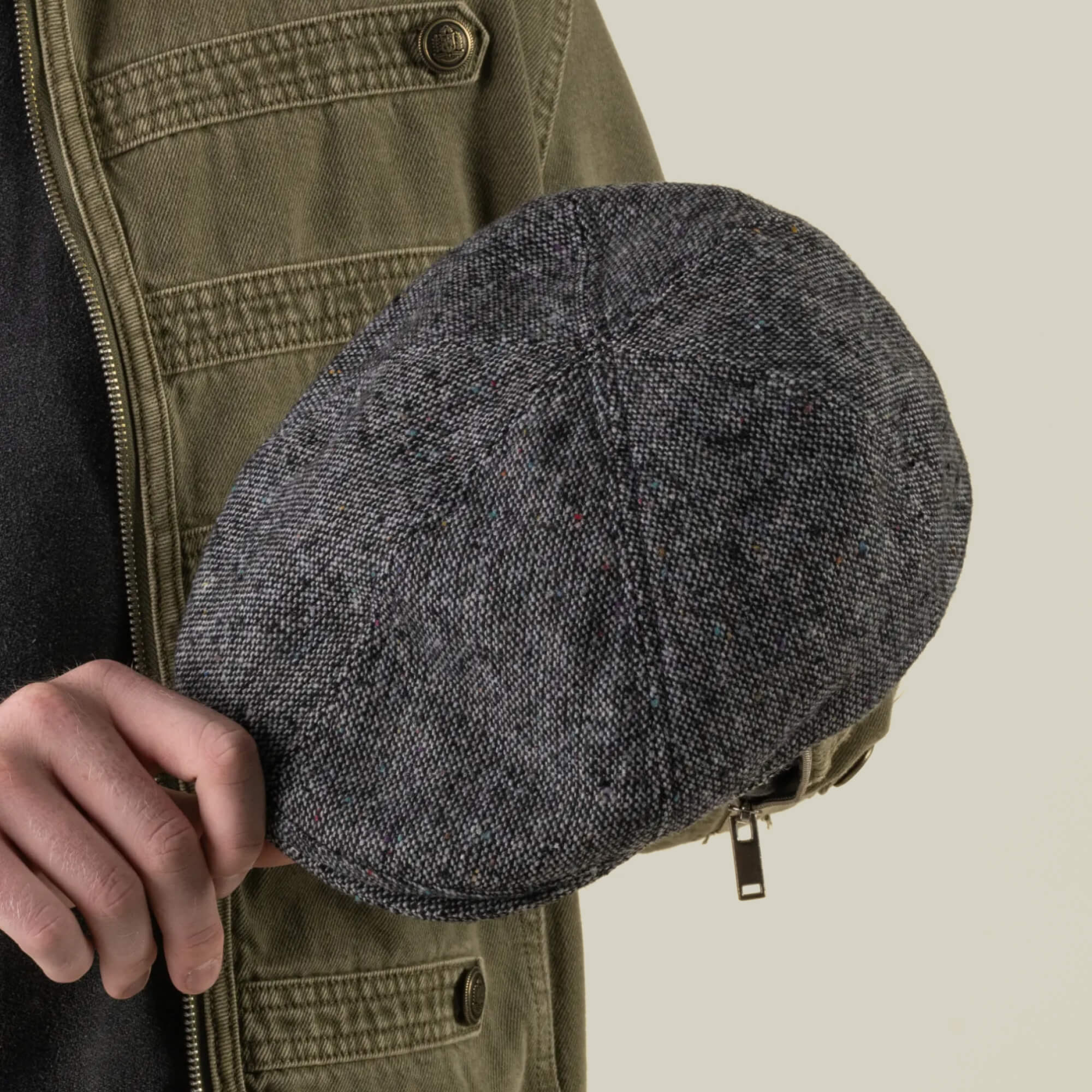 A person holds the Goorin Bros. Newlands digital gray flat cap in their right hand, wearing an olive green jacket over a black shirt against a neutral background.