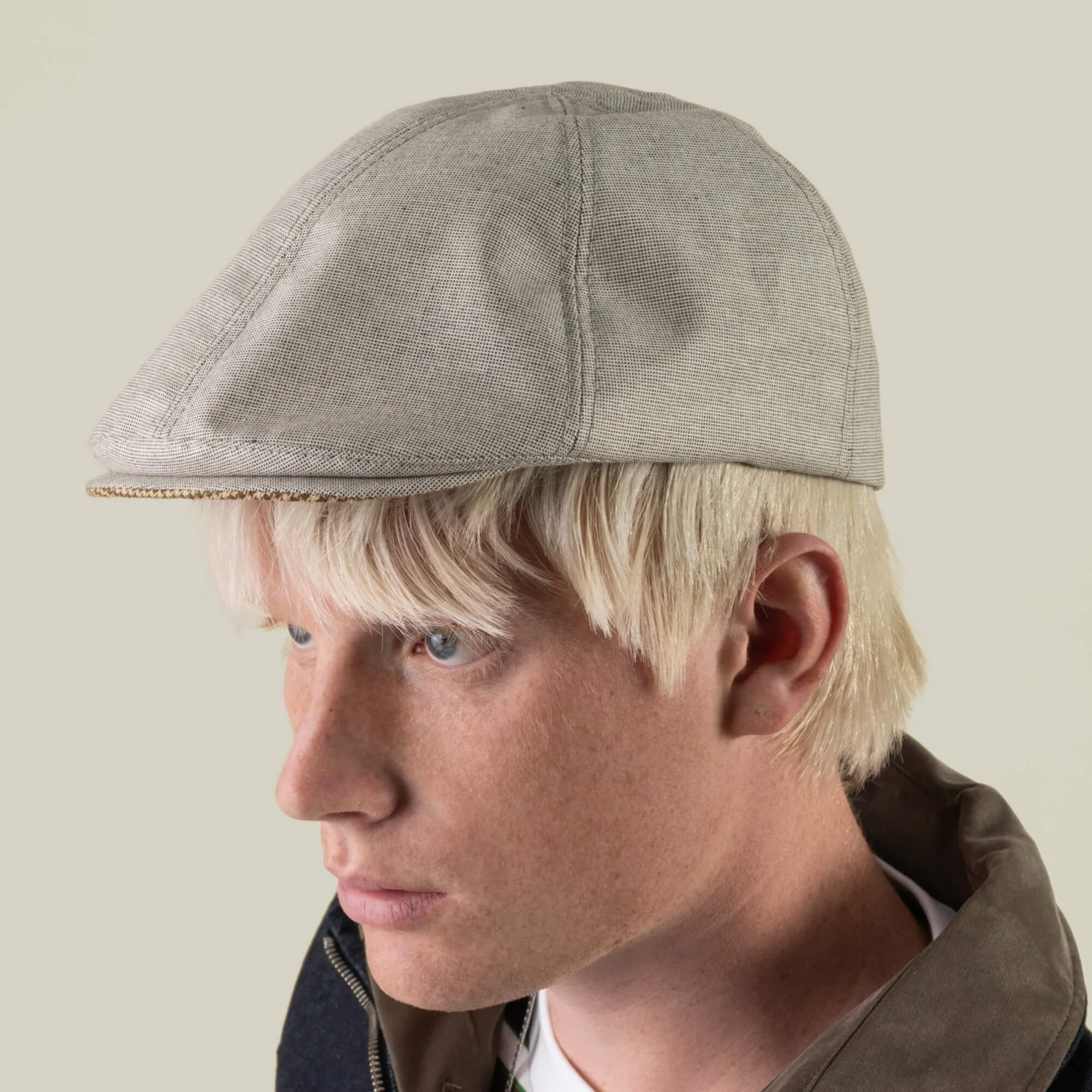 A person with light blond hair is shown in profile against a plain background, wearing the Goorin Bros. Mile Rock—a hand cut and sewn, light gray 6 Panel Ivy flat cap made in the USA—and a dark jacket.