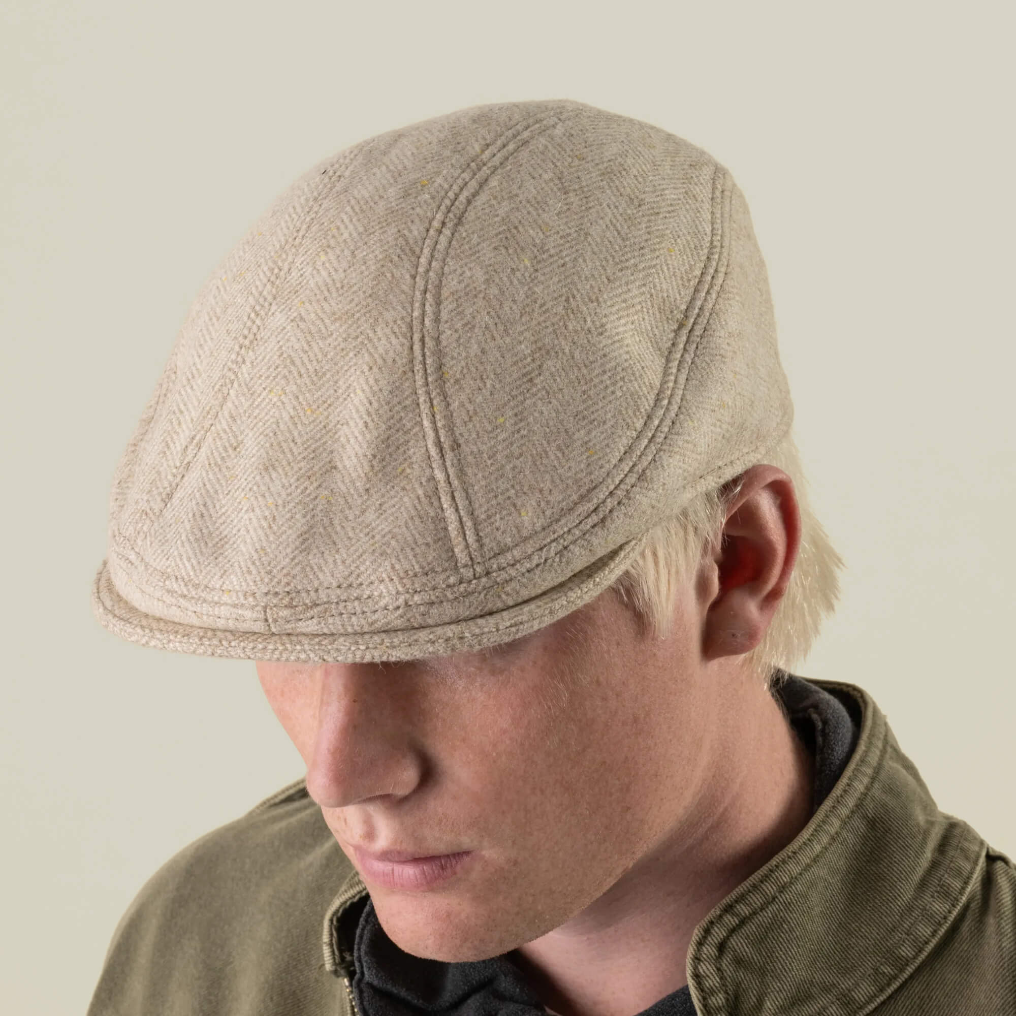 A person with light hair wears the Iris Fields beige flat cap by Goorin Bros.—a chalk cream headwear piece, hand-cut in the USA from 100% wool—paired with a green jacket and looking downward against a neutral background.