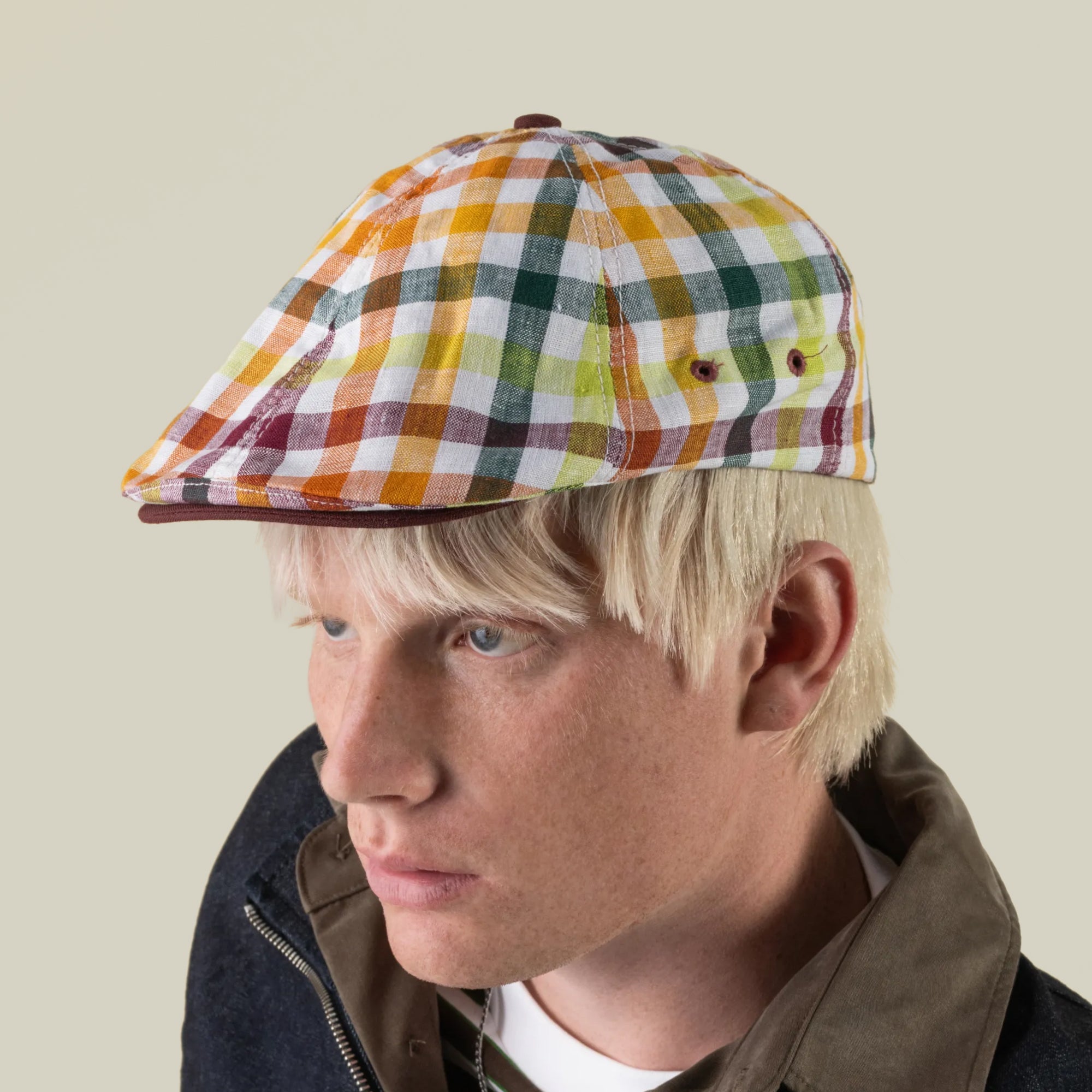A person with blond hair wears the “Point Lobos” 6 Panel Ivy flat cap by Goorin Bros., handmade in the USA from a breathable cotton blend in a multicolored plaid pattern, paired with a dark jacket, looking slightly to the left against a plain light background.