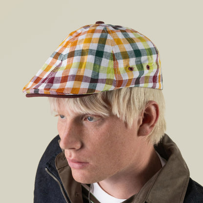 A person with blond hair wears the “Point Lobos” 6 Panel Ivy flat cap by Goorin Bros., handmade in the USA from a breathable cotton blend in a multicolored plaid pattern, paired with a dark jacket, looking slightly to the left against a plain light background.