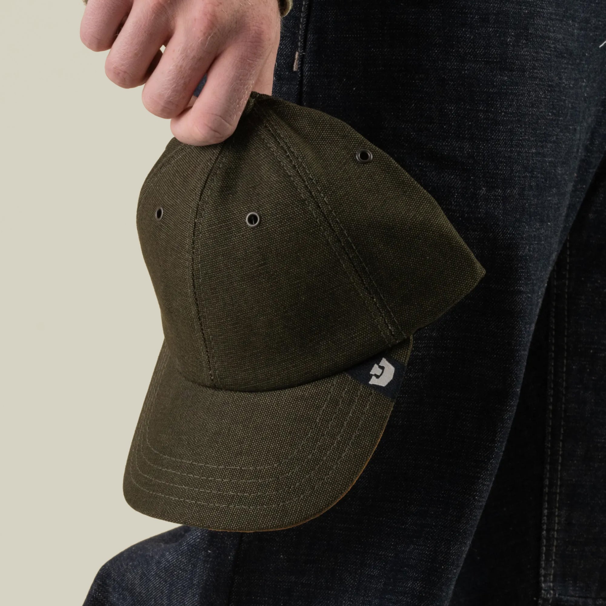 A person holds the Goorin Bros. Mineral Road cap—an olive green, mixed-material baseball cap made in the USA—by the top; the cap features a small tag on the brim, and the person is wearing dark jeans.