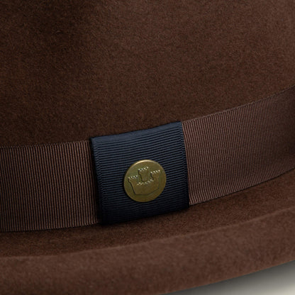Close-up of the Colonel Pierce hat by Goorin Bros.—a brown felt design with teardrop crown, brown grosgrain ribbon band, and a round gold emblem featuring a stylized hat logo.