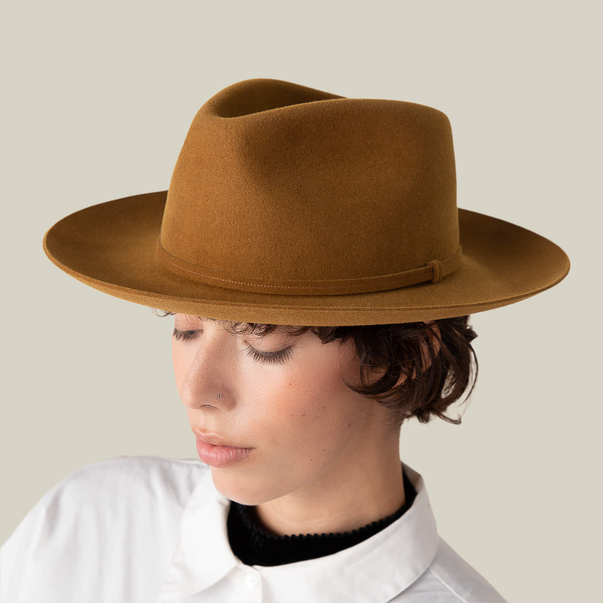 A person with short brown hair wears the Goorin Bros. Night Hawk brown fedora, featuring a wide brim and teardrop crown, paired with a white collared shirt, looking downward against a plain background.