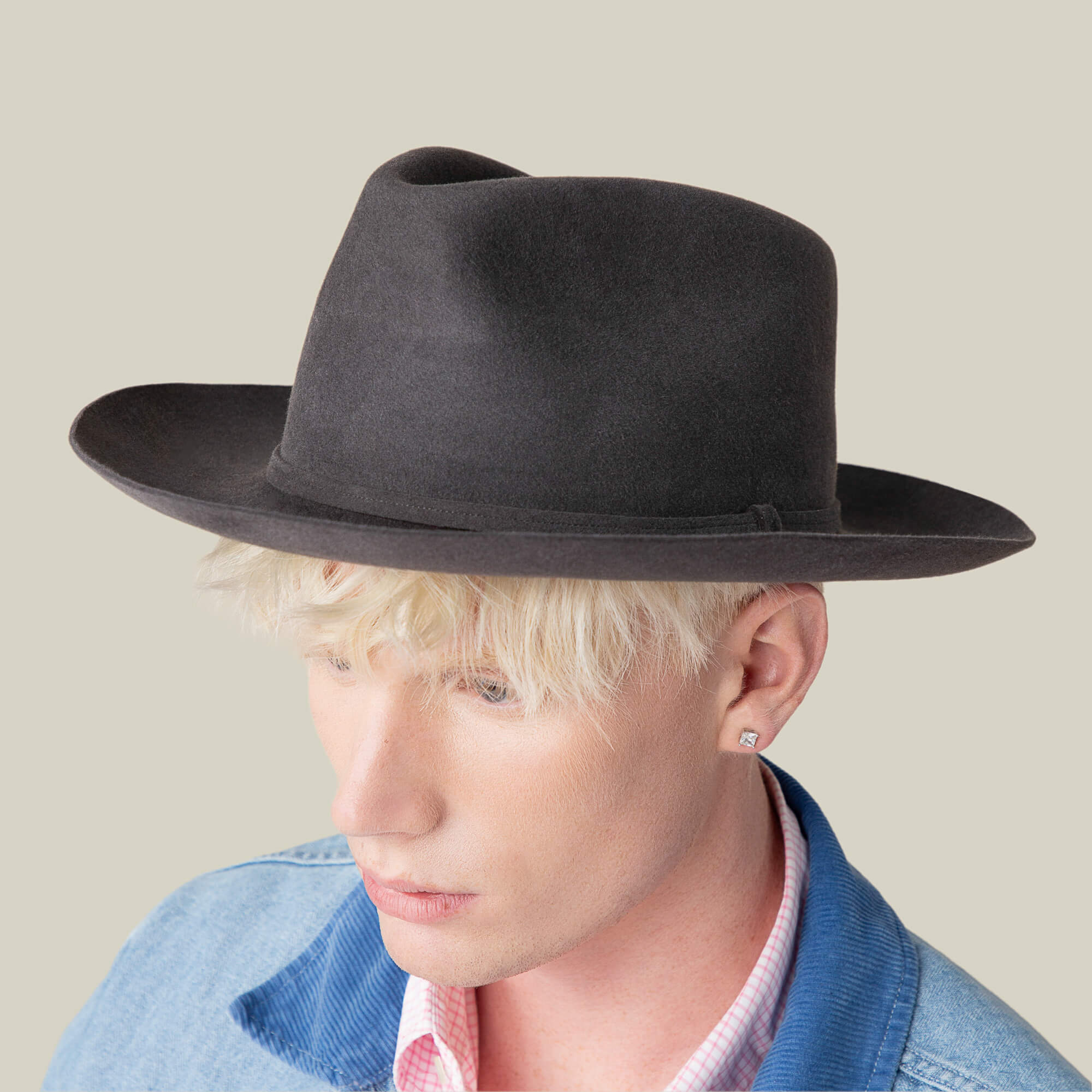 A person with light blond hair faces slightly downward, wearing the Goorin Bros. Night Hawk black fedora hat with a teardrop crown and wide brim, paired with a light blue jacket and pink collared shirt.