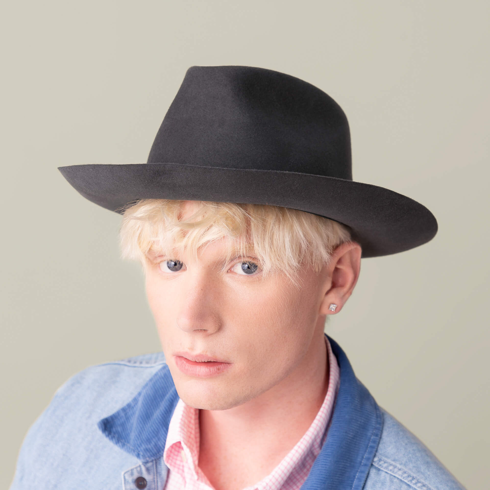 A person with light blonde hair wears the Goorin Bros. Night Hawk black wide-brimmed fedora, a denim jacket, and a stud earring while looking at the camera against a plain background.