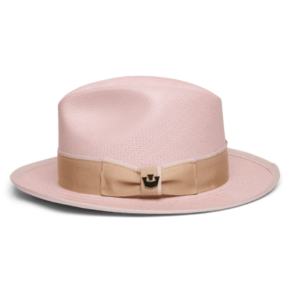 The Hartman by Goorin Bros. is a light pink fedora made from 100% toquilla, featuring a wide beige grosgrain band with bow, metallic accent, and contrast edge binding, displayed on a plain white background.