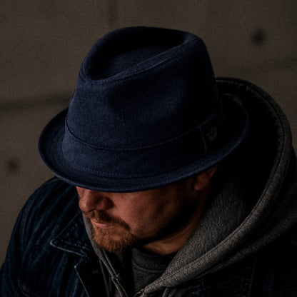 A man wearing Goorin Bros. Charlie Pants, a navy blue linen fedora, a gray hoodie, and a denim jacket looks downward in a dimly lit setting.