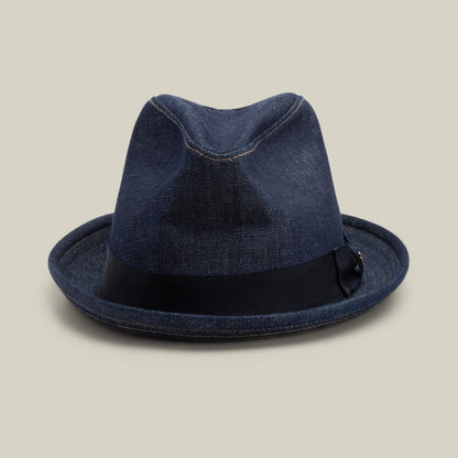 The Goorin Bros. Noisy Big Stepper fedora in indigo denim features a sharp crown and black ribbon band, showcased on a plain light beige background.
